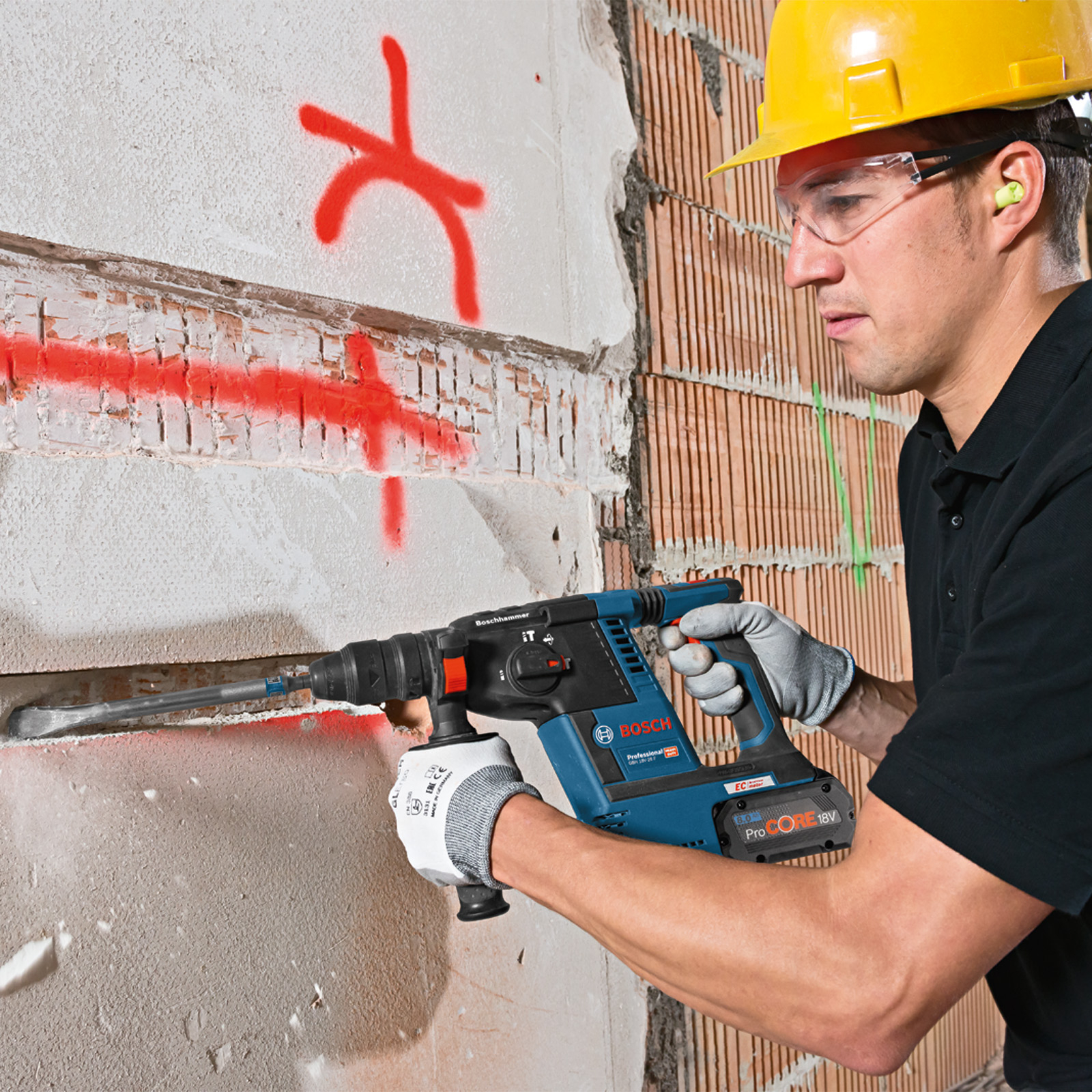 Ein Mann mit gelbem Helm bohrt mit einem Bosch-Hammer in eine Wand. Rote Markierungen sind an der Wand. Der Mann trägt Brille und Handschuhe.