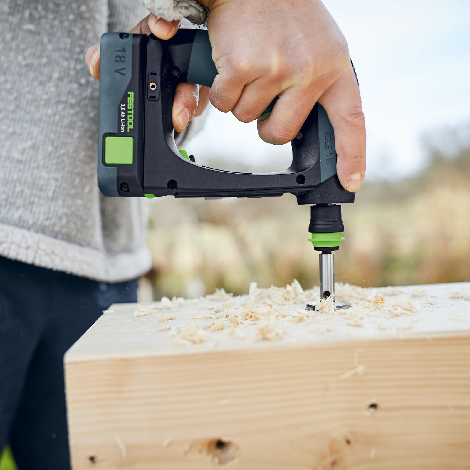 Eine Person bohrt mit einem schwarz-grünen Bohrer in einen Holzblock. Holzspäne fliegen. Der Bohrer ist ein Festool CXS 18.