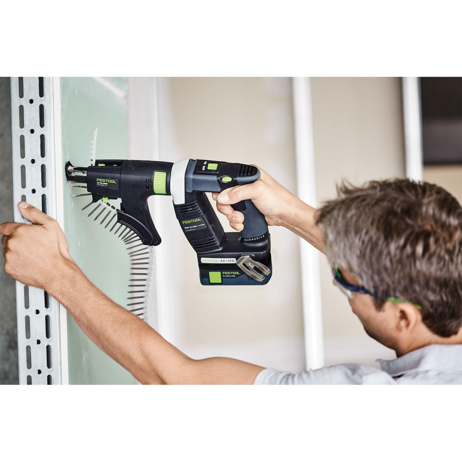 Un uomo usa un avvitatore Festool nero e verde per installare cartongesso. Indossa occhiali protettivi e tiene un montante metallico.