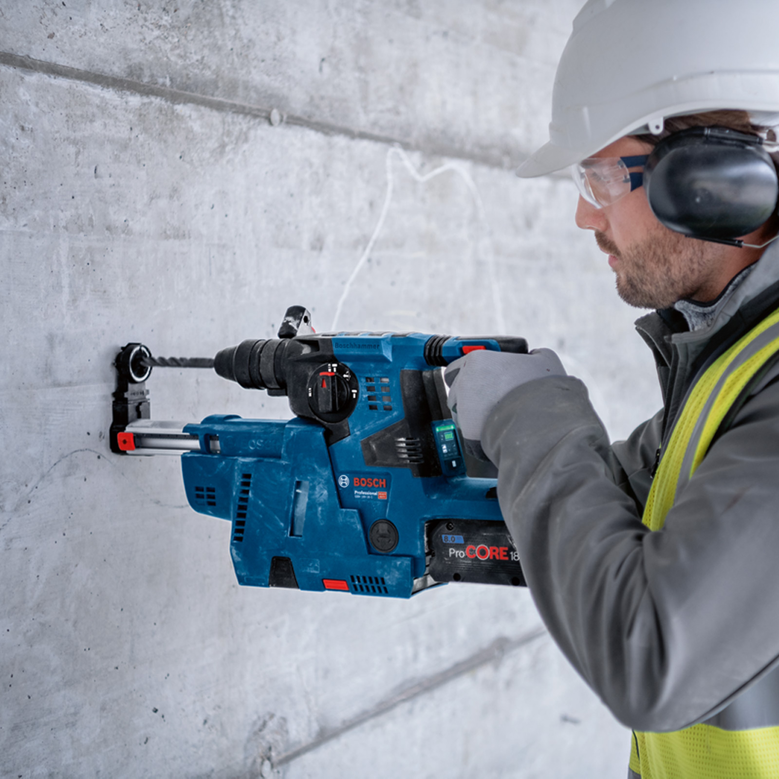 Robotnik budowlany wierci w betonowej ścianie niebieską wiertarką udarową Bosch, w odzieży ochronnej.