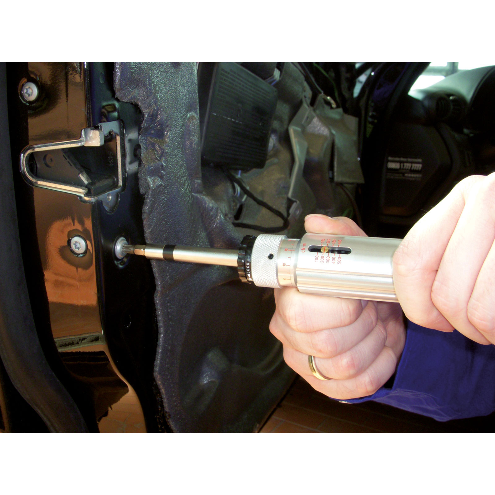 Eine Hand verwendet einen silbernen Drehmomentschlüssel, um eine Schraube am Türrahmen eines Autos festzuziehen. Die Tür ist schwarz, der Innenraum ist sichtbar.