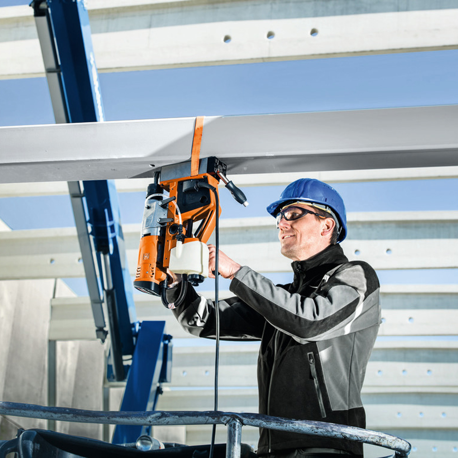 Ein Arbeiter bohrt mit einer orangefarbenen Bohrmaschine in einen Betonbalken. Er trägt einen Helm und eine Schutzbrille. Der Himmel ist blau.