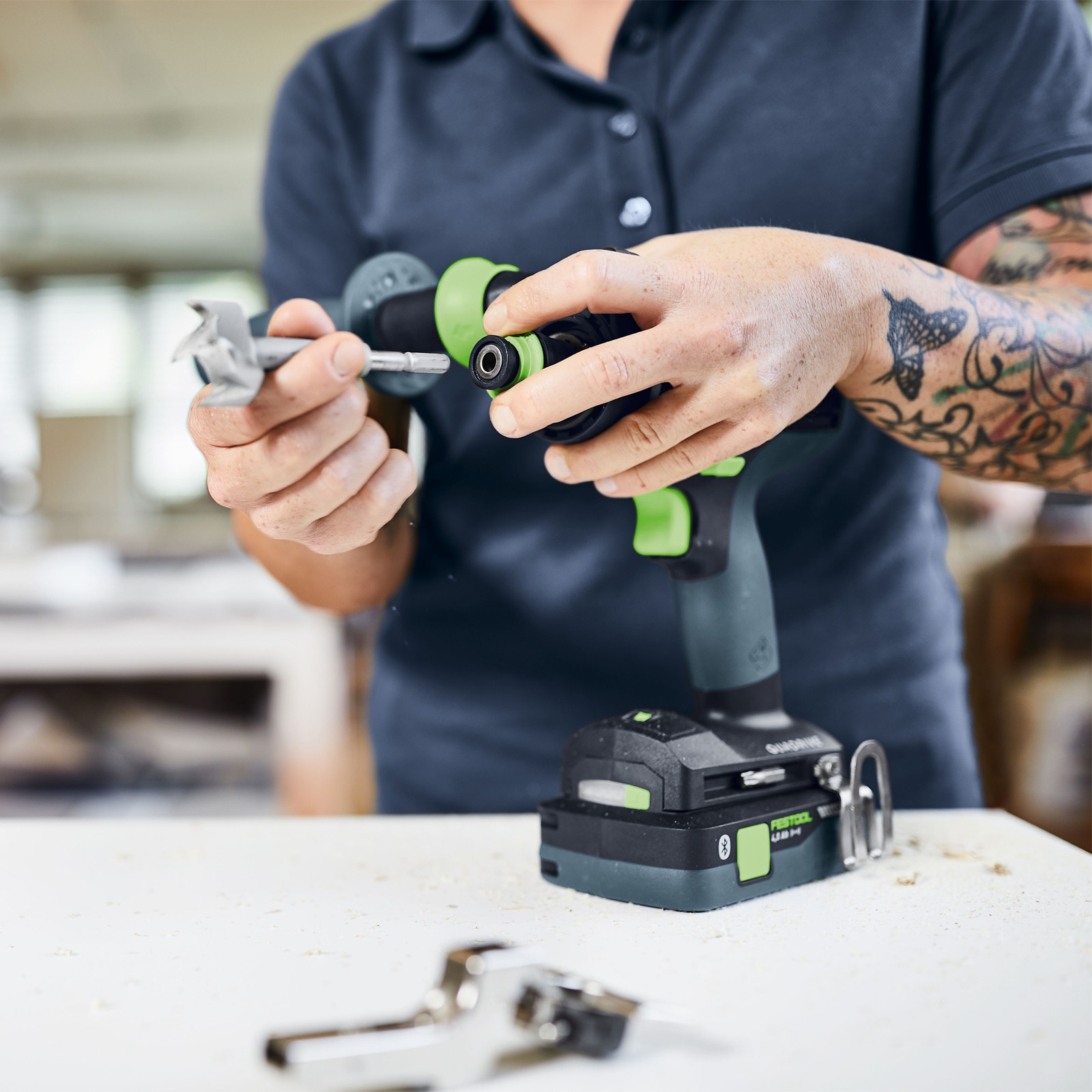 Eine Person montiert einen Bohrer. Sie halten einen Bohrer und einen Bohrer. Die Person hat ein Tattoo auf dem Arm. Ein Metallscharnier liegt auf dem Tisch.
