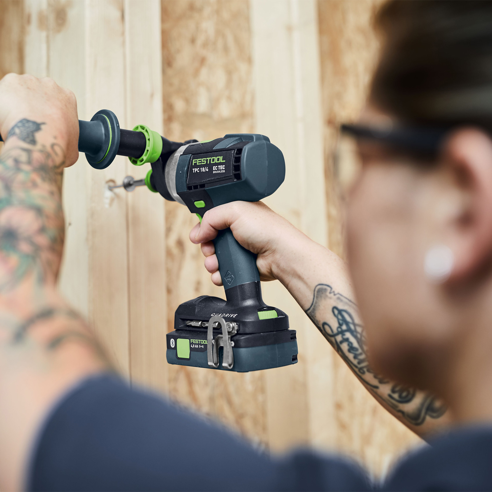 Eine Person bohrt mit einem schwarz-grünen Akkuschrauber in eine Holzwand. Die Person hat Tattoos auf dem Arm.