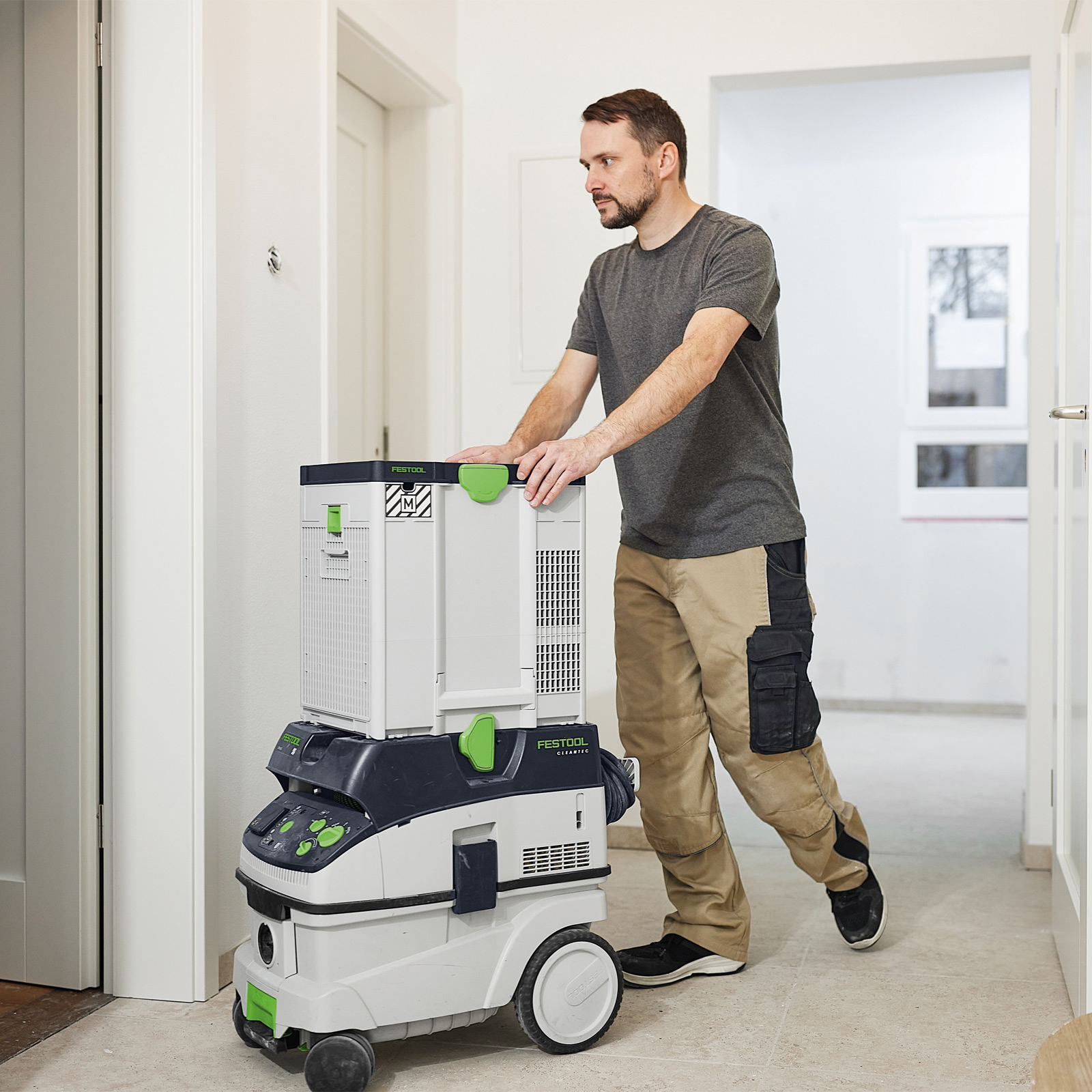 Ein Mann in Arbeitskleidung schiebt einen Festool-Staubsauger einen Flur entlang. Der Staubsauger ist weiß, schwarz und grün.