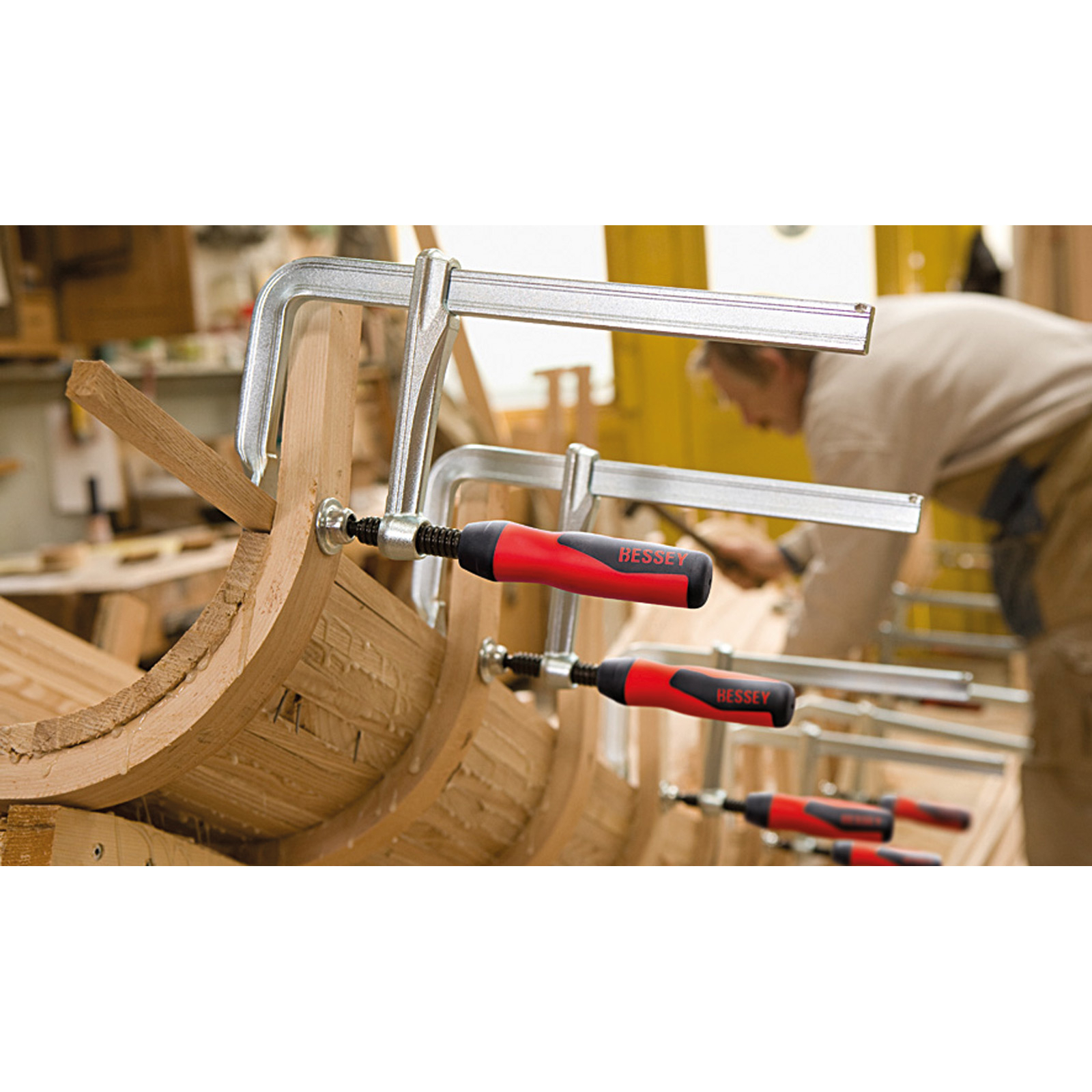 Stolarz używa zacisków do łączenia zakrzywionych kawałków drewna. Widoczne są zaciski z czerwonymi uchwytami. Osoba pracuje w tle.