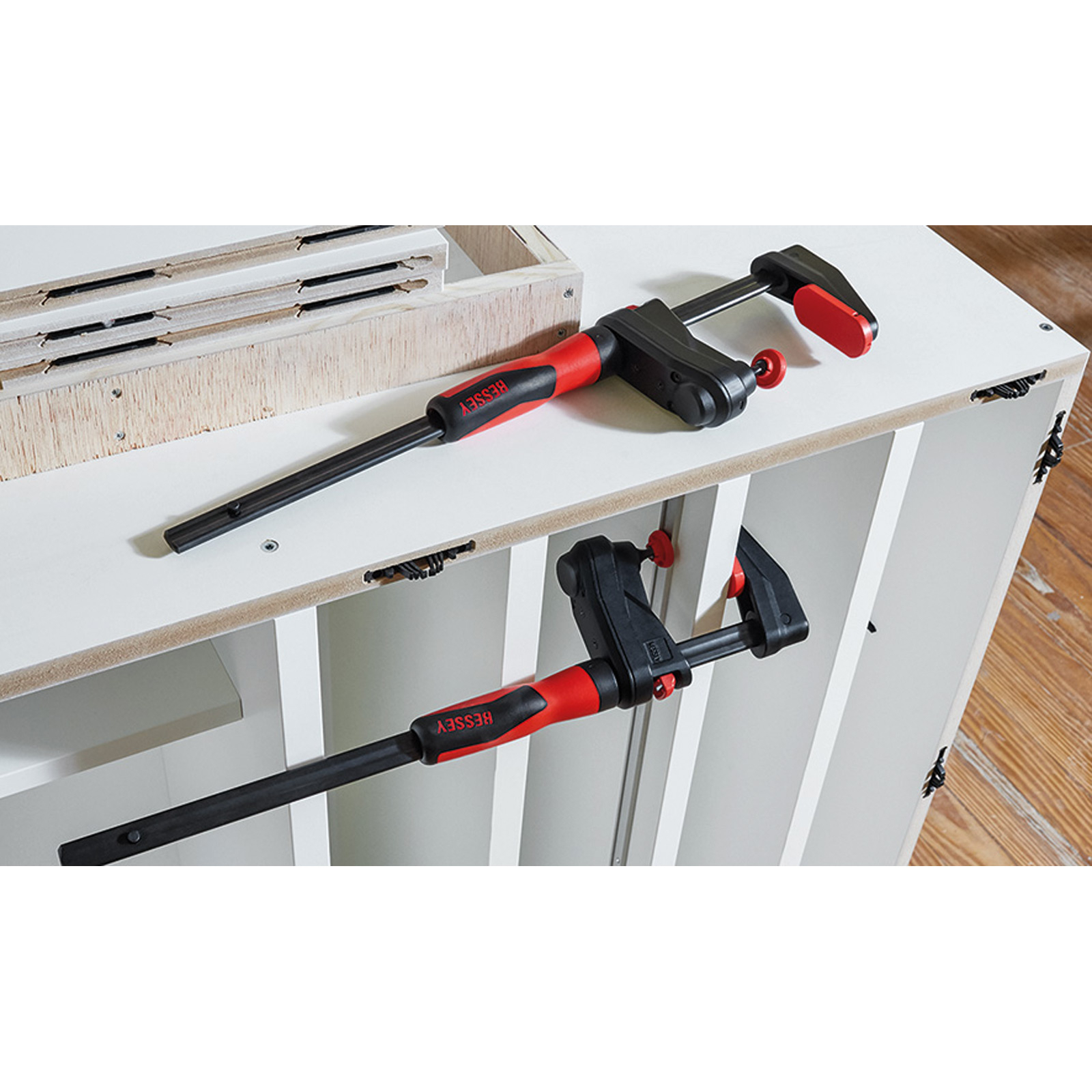 Due morsetti su un mobile bianco. Un morsetto tiene un pezzo di legno. Manici rossi e neri. Cornice in legno sullo sfondo.