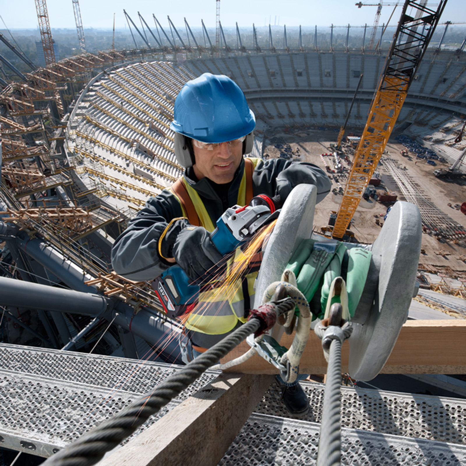 Robot budowlany w niebieskim kasku używa szlifierki do metalowej konstrukcji. Iskry lecą. Tło: stadion w budowie.