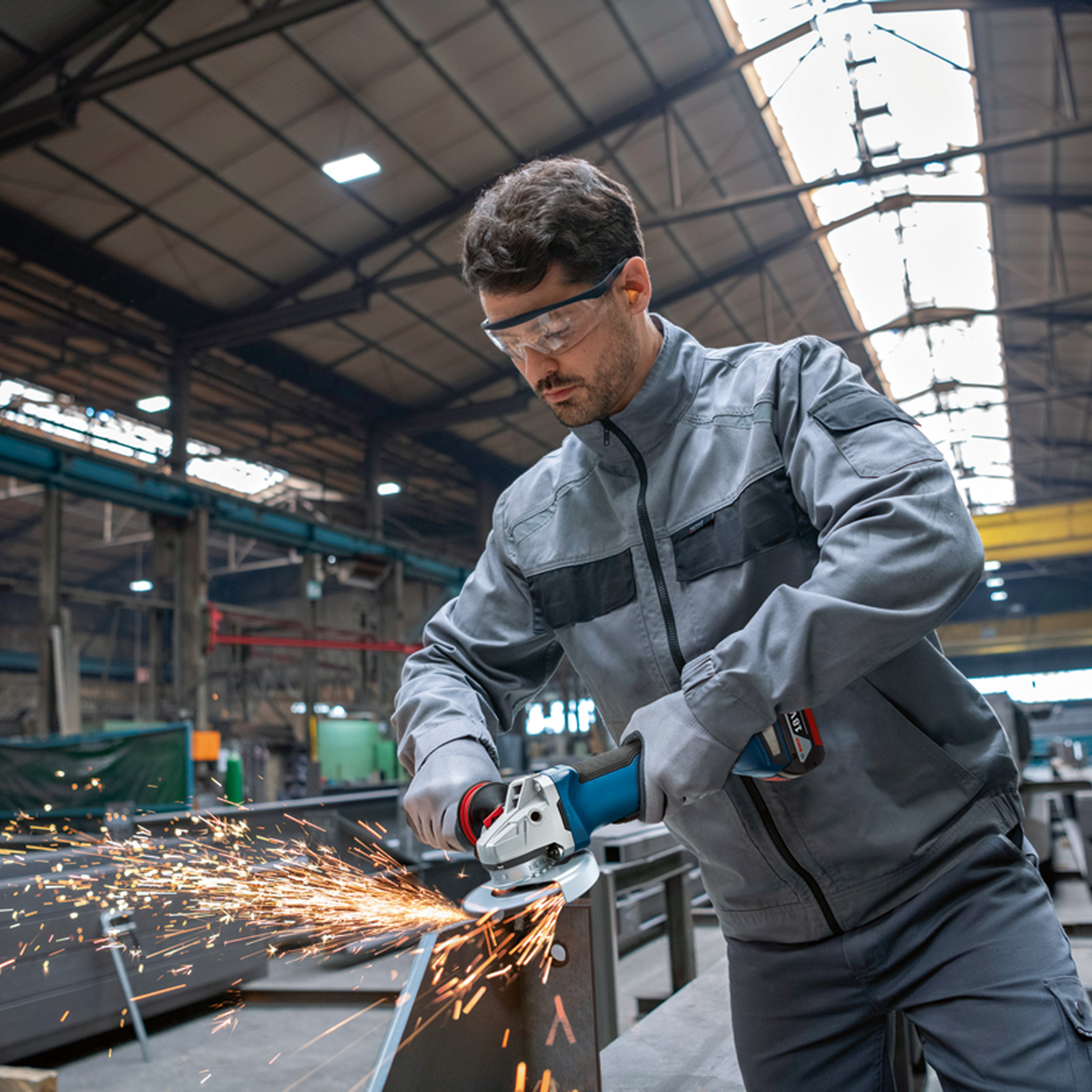 Mężczyzna w szarym ubraniu roboczym używa szlifierki kątowej, lecą iskry. Przemysłowe otoczenie.