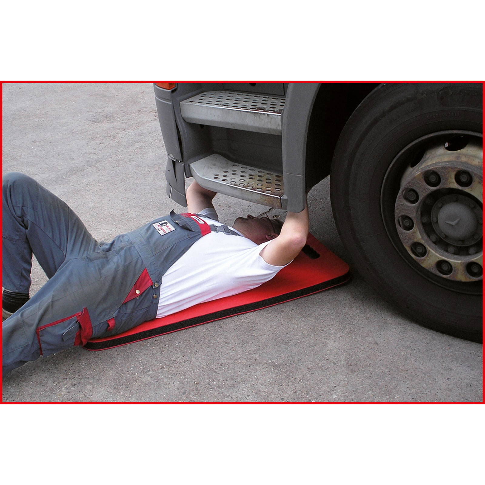 Un meccanico è sdraiato sotto un camion, su un cuscinetto rosso. L'uomo indossa una tuta e una camicia bianca. Il camion ha una gomma.