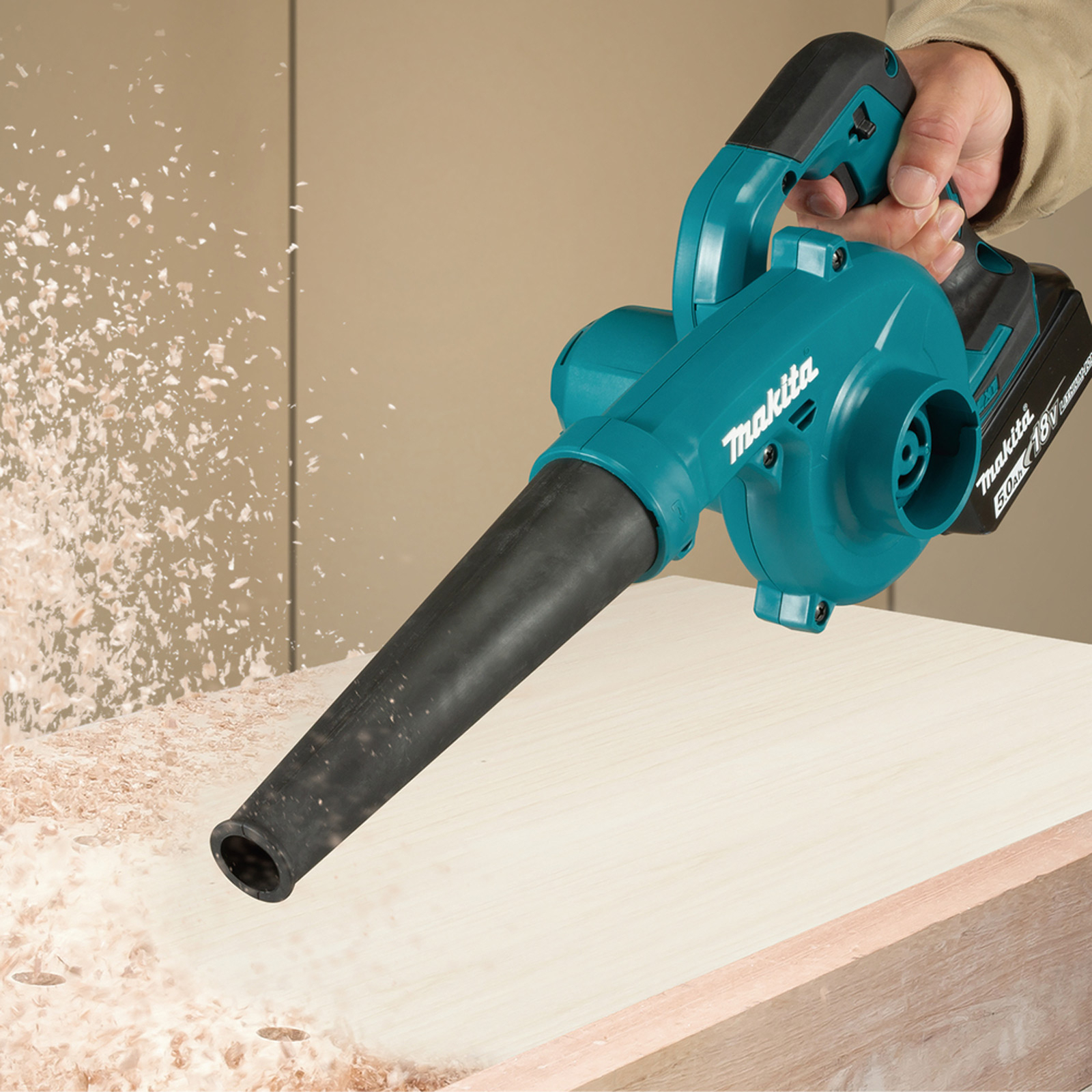 Ein Makita-Laubbläser, türkis und schwarz, bläst Holzspäne von einer hellen Holzoberfläche. Eine Hand hält das Werkzeug.