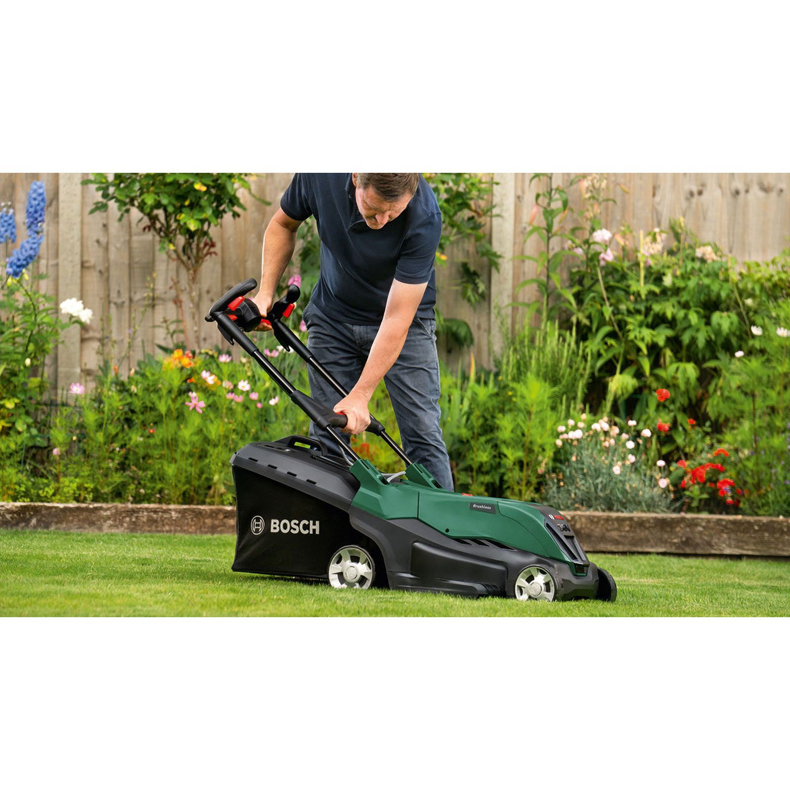 Un hombre corta el césped con un cortacésped Bosch verde y negro en un jardín. Flores y una cerca en el fondo.