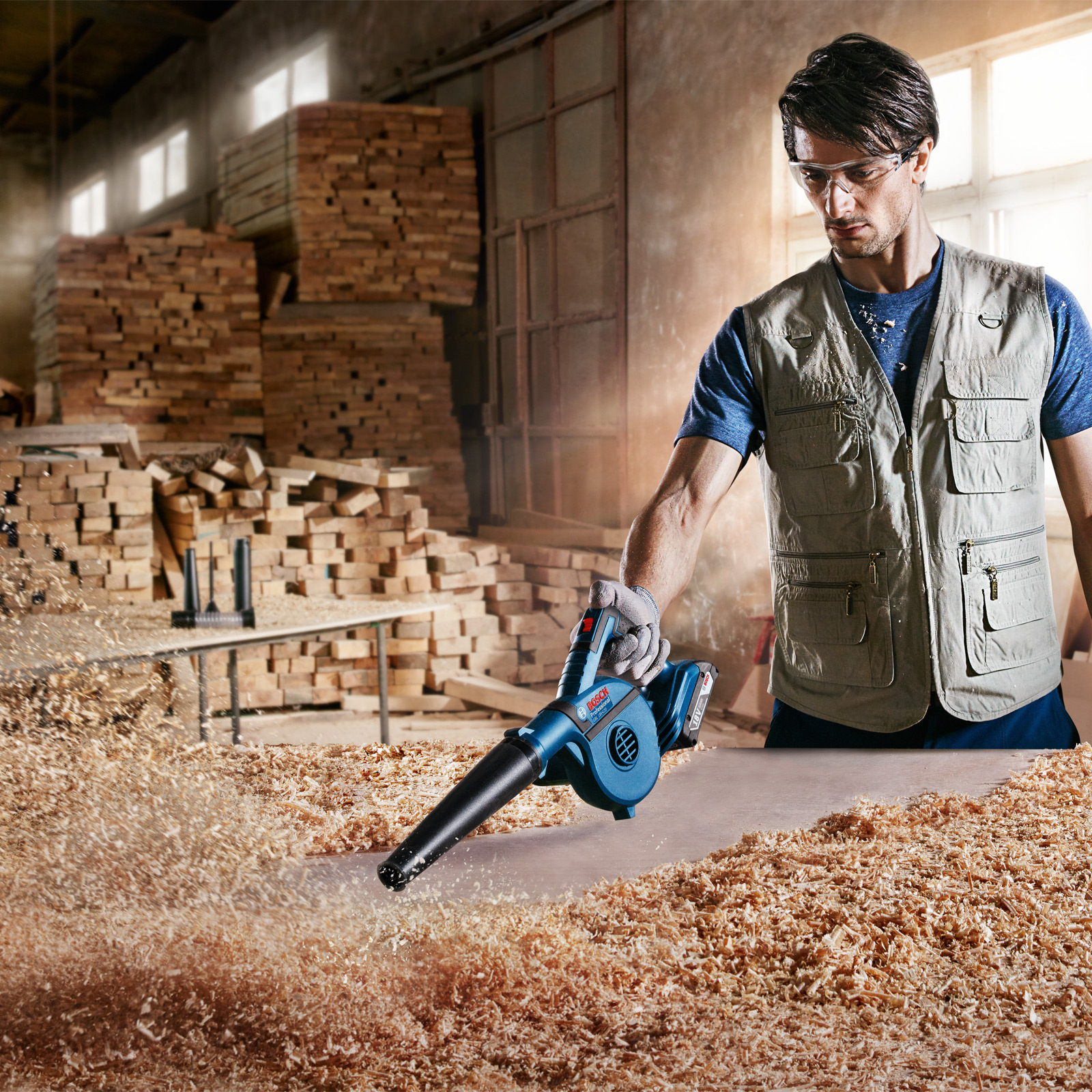 Un hombre usa un soplador para limpiar virutas de madera. Usa gafas de seguridad y chaleco. Hay pilas de madera detrás.