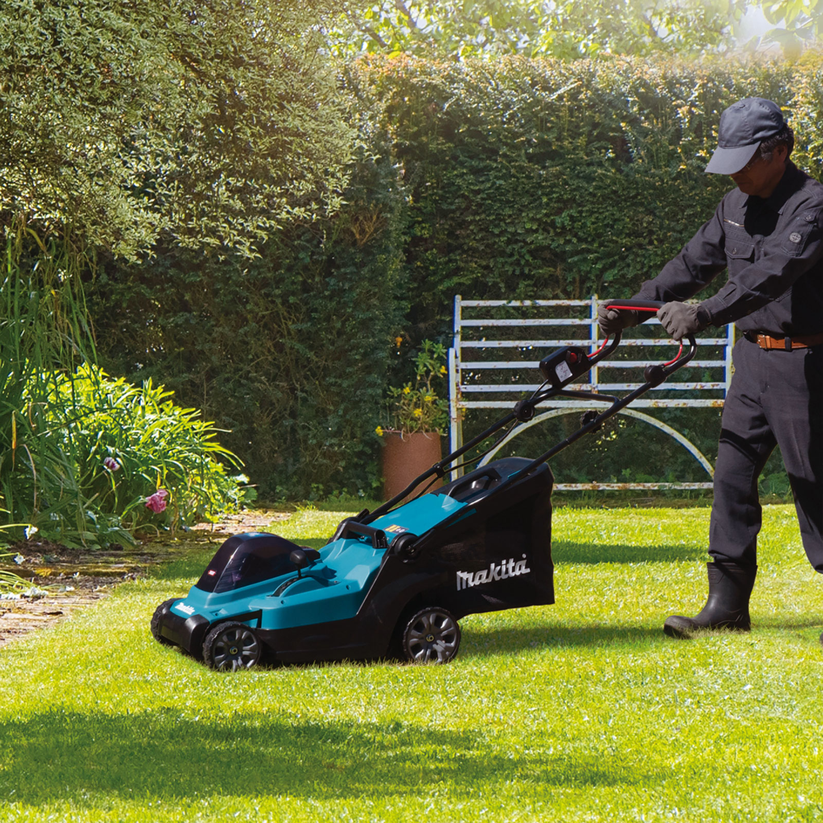 Ein Mann in schwarzer Arbeitskleidung mäht einen Rasen mit einem Makita-Rasenmäher. Grünes Gras, Hecken und ein weißer Zaun im Hintergrund.