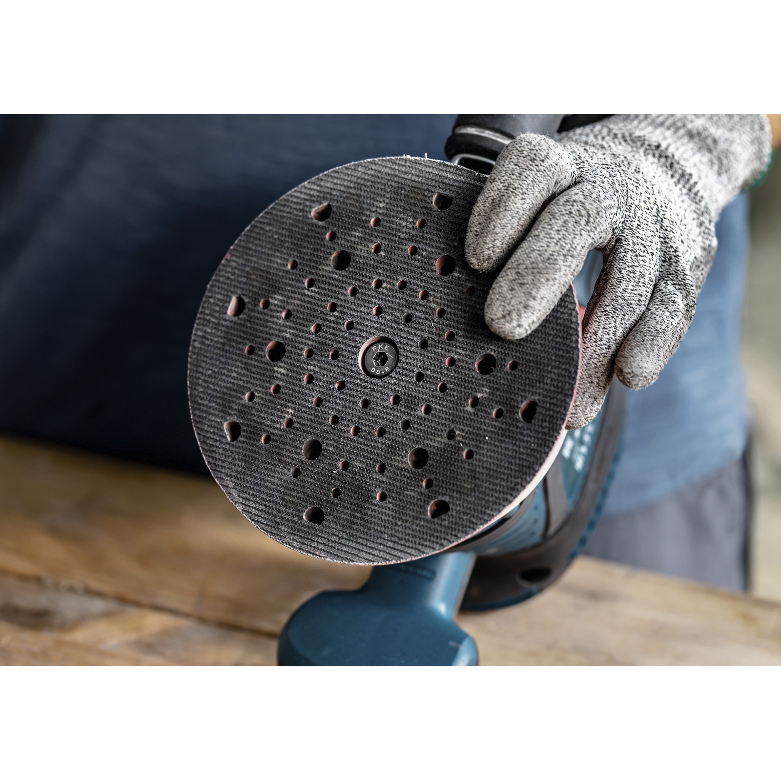 Una persona con guanto tiene in mano un disco abrasivo. Il disco è nero con fori e la levigatrice è blu.