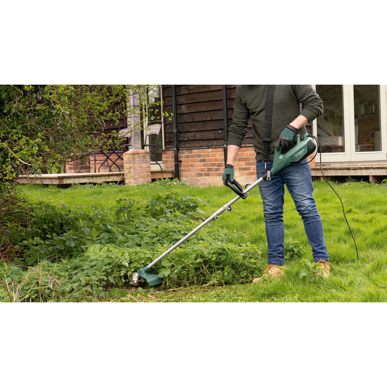 Ein Mann benutzt einen grünen und schwarzen Rasentrimmer. Der Mann trägt blaue Jeans, grüne Handschuhe und ein grünes Hemd. Der Hintergrund ist ein Haus und Gras.