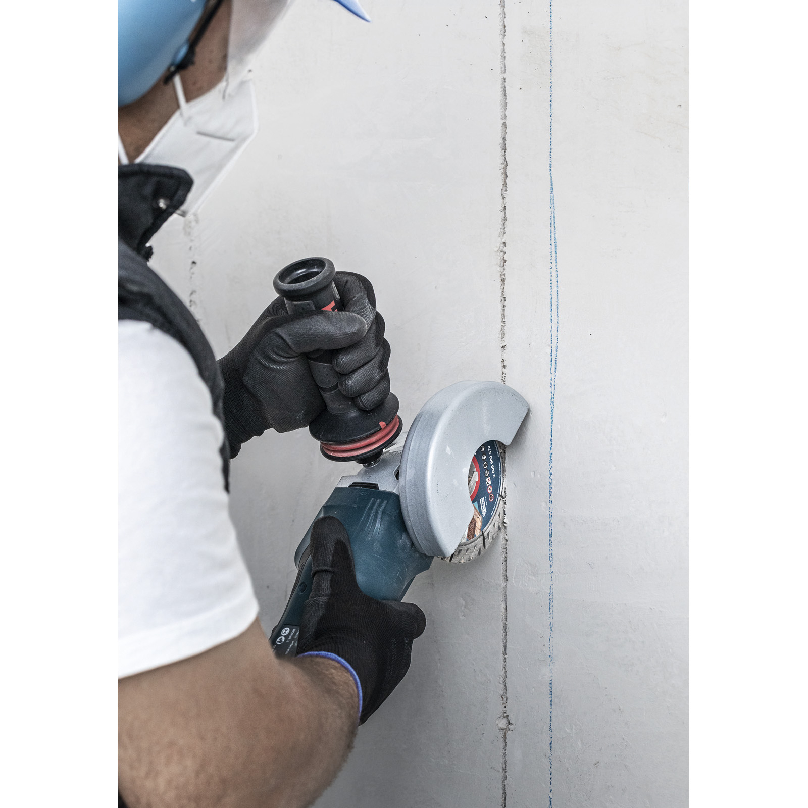 Ein Arbeiter schneidet mit einem Winkelschleifer eine Linie in eine Betonmauer. Er trägt Handschuhe, eine Maske und eine Schutzbrille.
