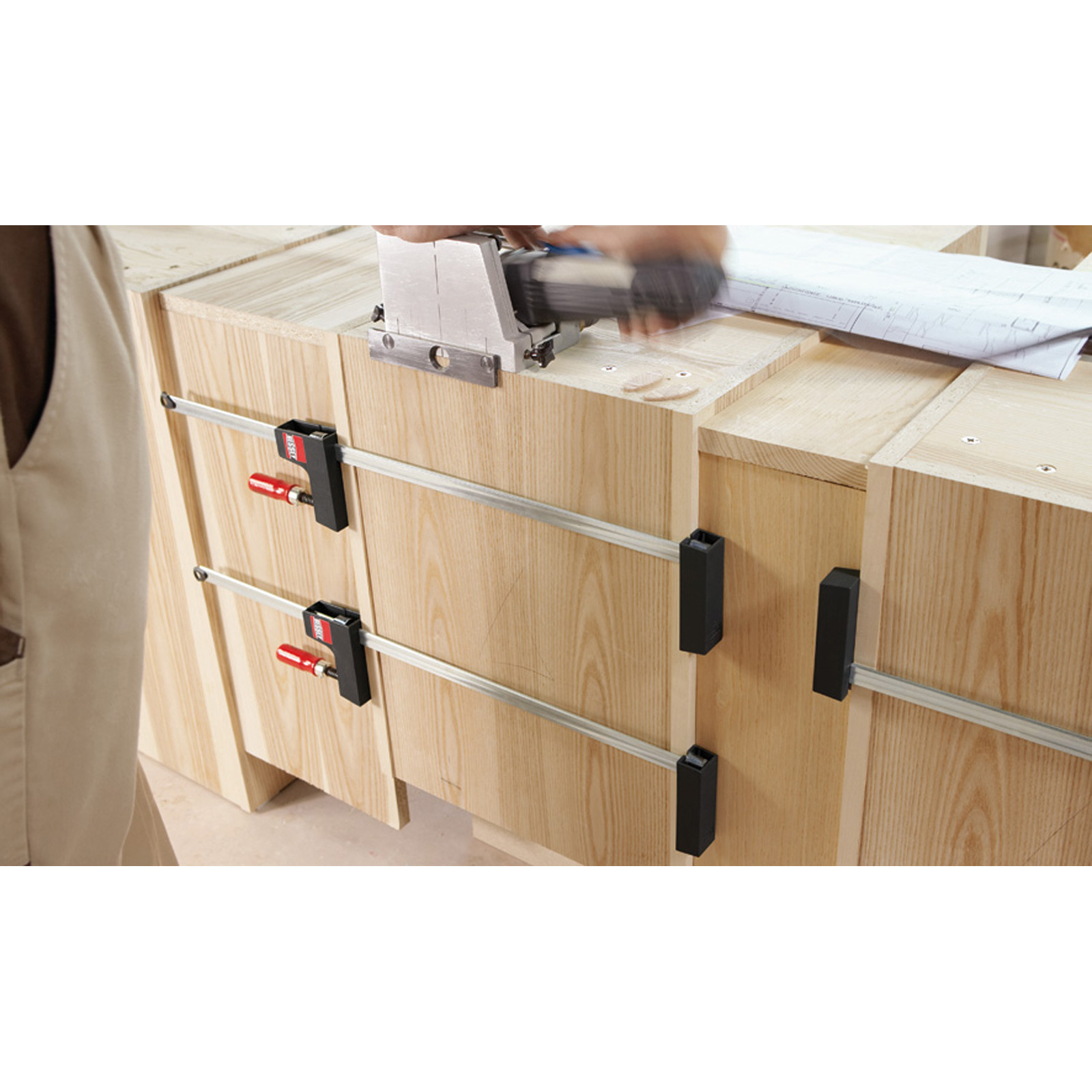 Ein Tischler verwendet eine Dübelfräse an einem Holzschrank. Klemmen sichern Teile. Baupläne liegen auf der Oberfläche.