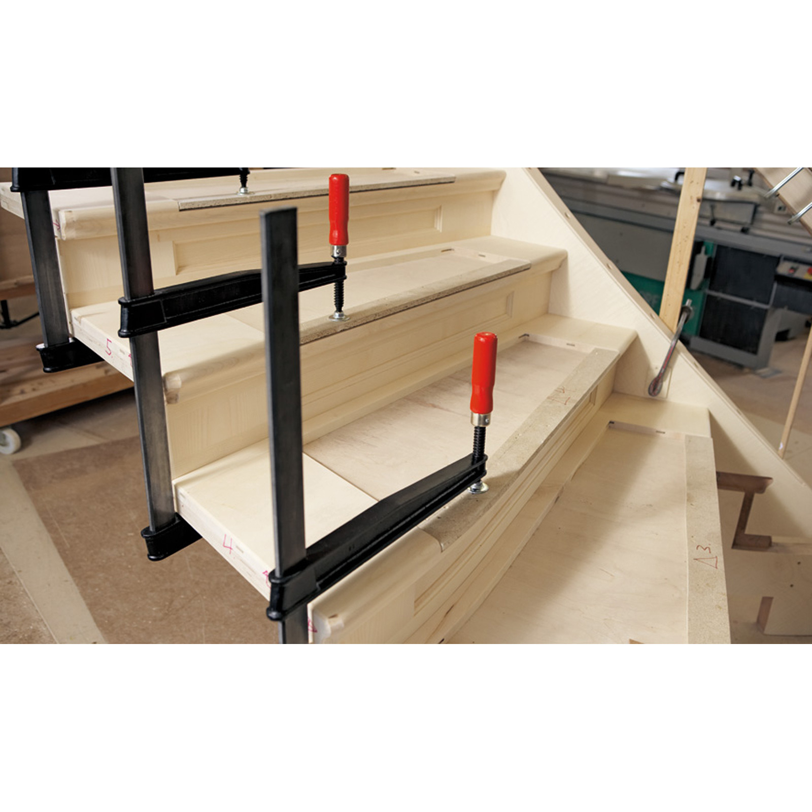 Drewniane schody w budowie z zaciskami. Widoczne czerwone uchwyty. Scena w warsztacie.