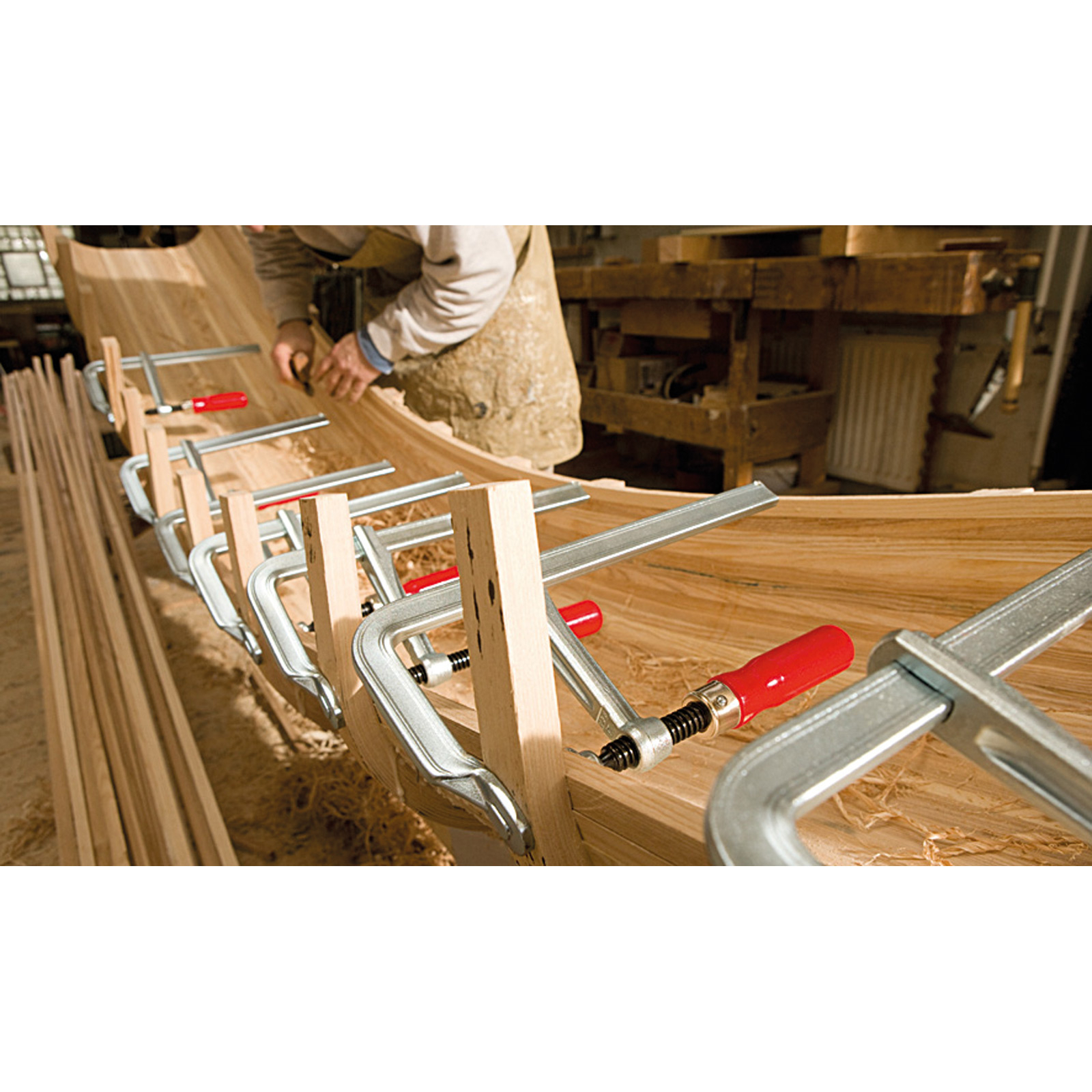 Ein Zimmermann verwendet Klemmen an einem Holzbootrahmen. Rotgriffige Klemmen halten Holzstücke zusammen. Sägemehl sichtbar.