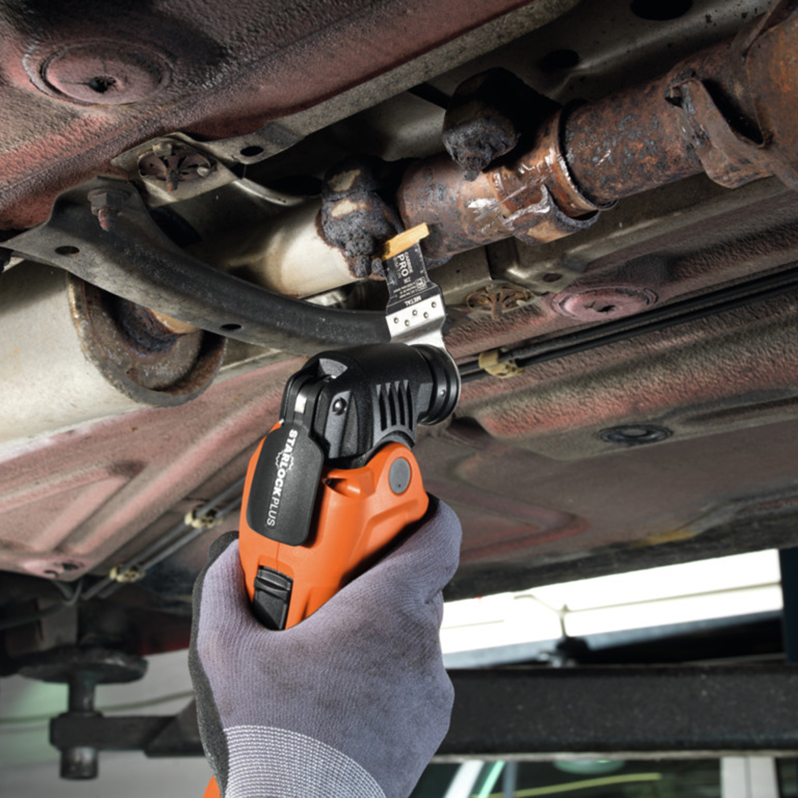 Eine Person verwendet ein orangefarbenes und schwarzes Oszillations-Multitool, um ein Autoauspuffrohr zu schneiden. Das Werkzeug wird von einer behandschuhten Hand gehalten.
