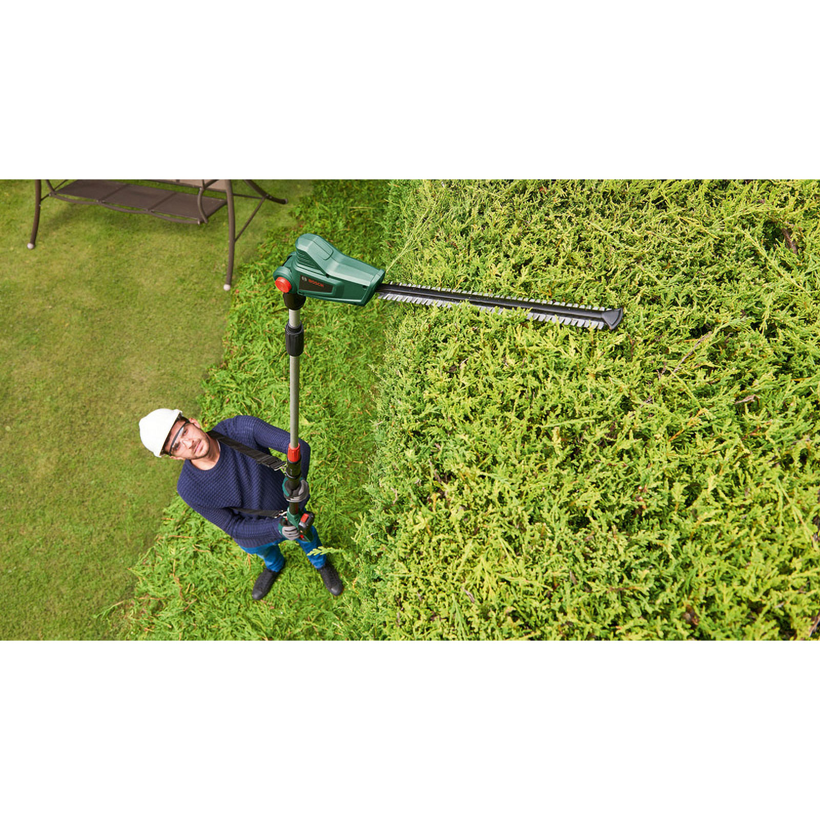 Mężczyzna przycina żywopłot zielono-czarną przycinarką. Ma na sobie kask i okulary ochronne. Zielona trawa i żywopłot w tle.