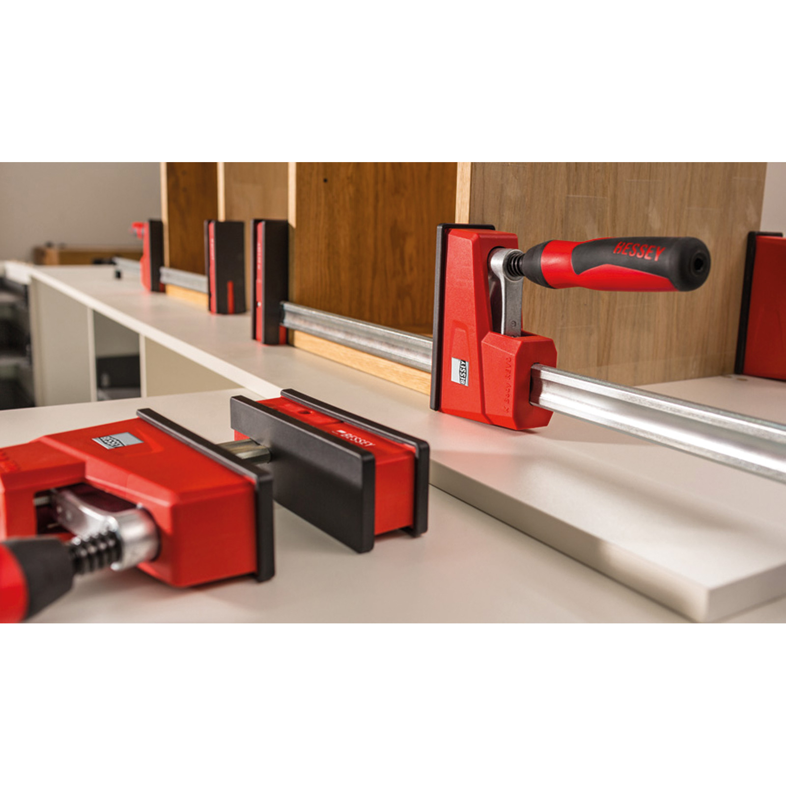 Rote BESSEY-Klemmen halten Holzplatten fest. Die Szene umfasst ein weißes Regal und einen Werkstatthintergrund.