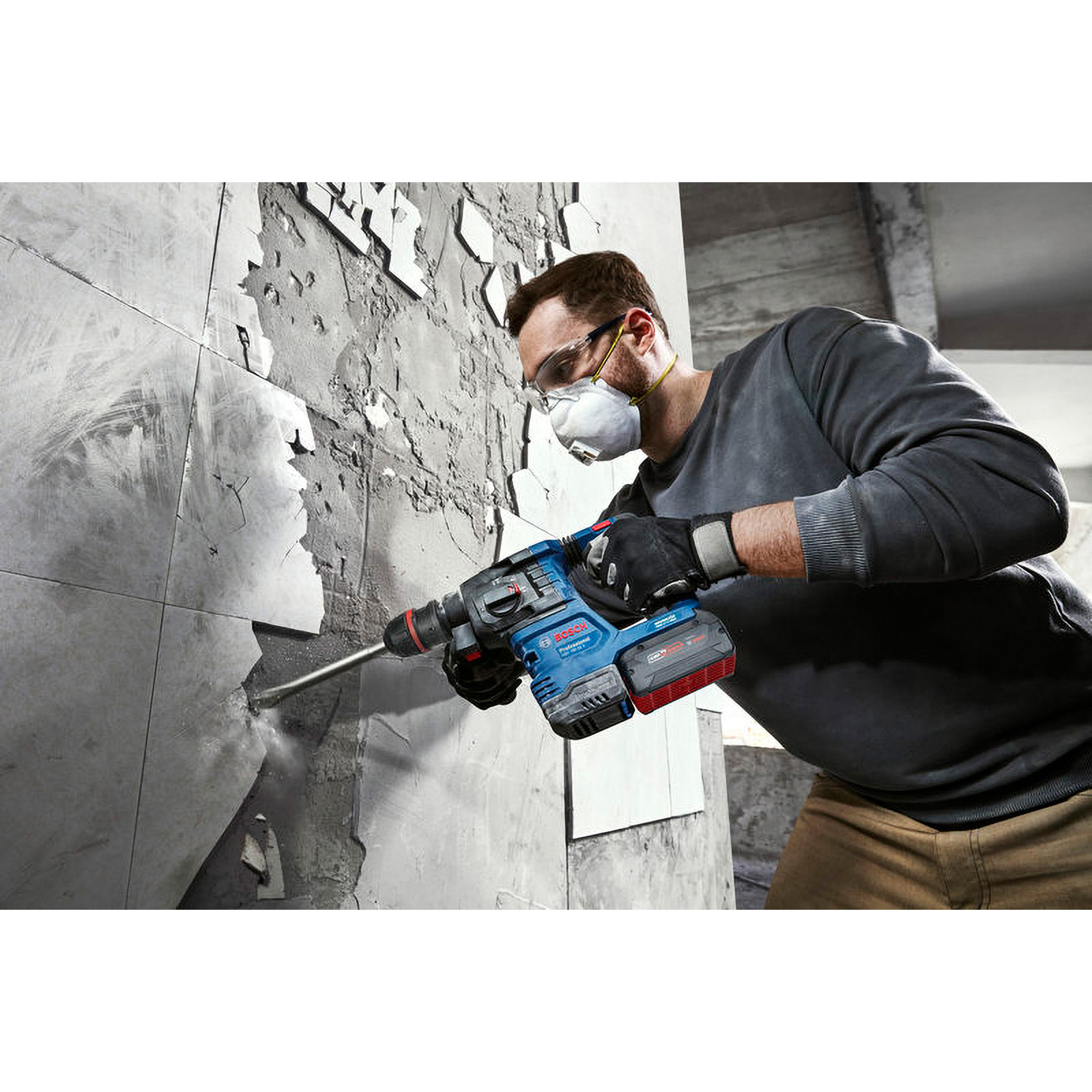 Ein Mann benutzt einen Bohrhammer, um eine Fliesenwand aufzubrechen. Er trägt eine Schutzbrille, eine Maske, Handschuhe und ein graues Hemd.