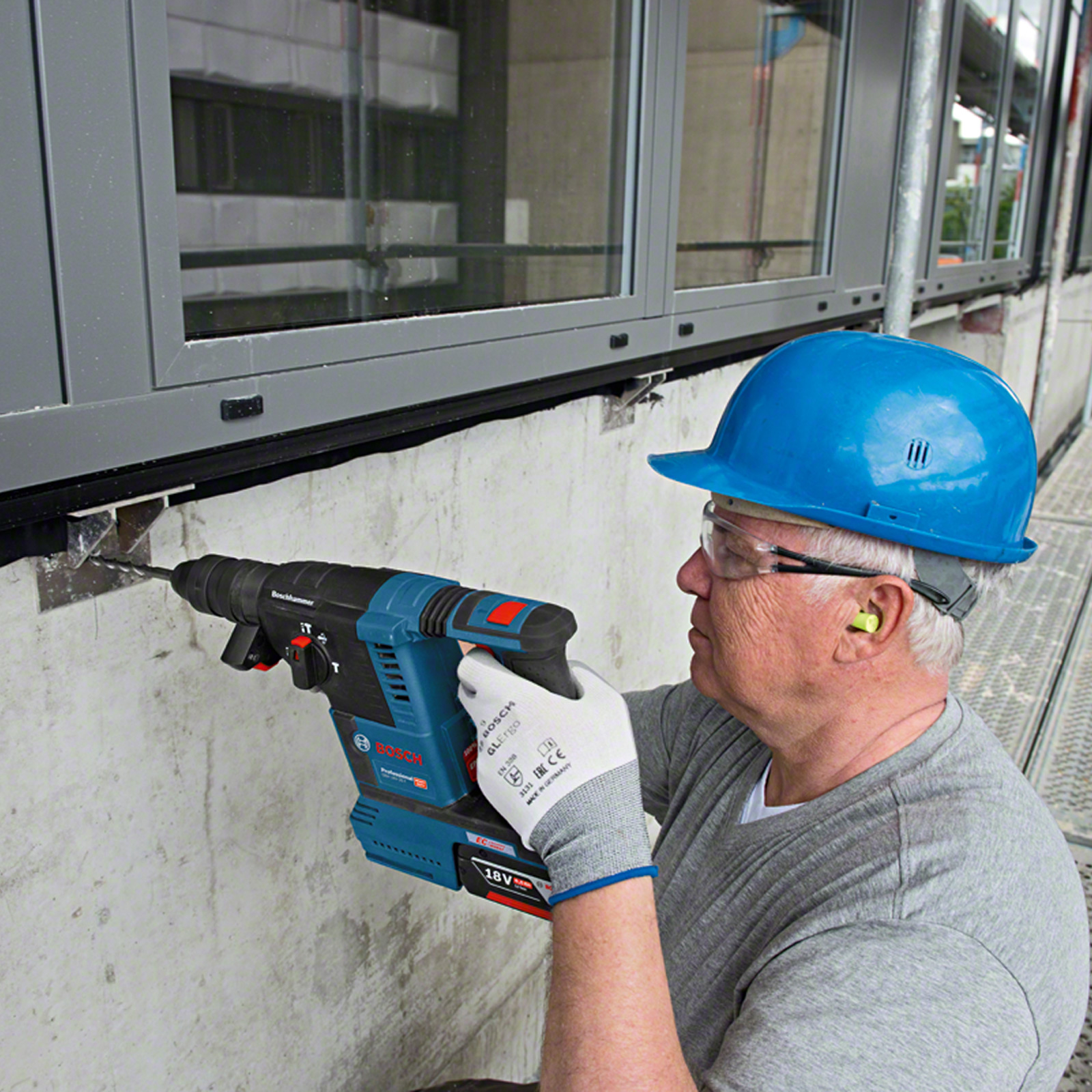 Mężczyzna wierci w betonowej ścianie niebieską wiertarką udarową Bosch. Ma niebieski kask, okulary ochronne i zatyczki.