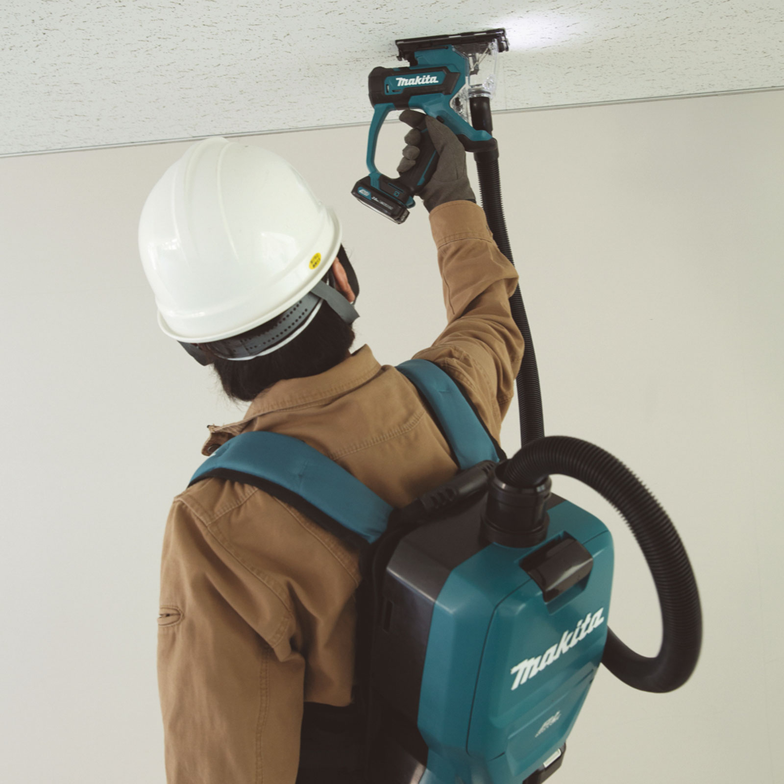 Un trabajador usa una herramienta Makita en un techo. Usa un casco y una aspiradora de mochila está conectada. La herramienta es verde azulado y negra.