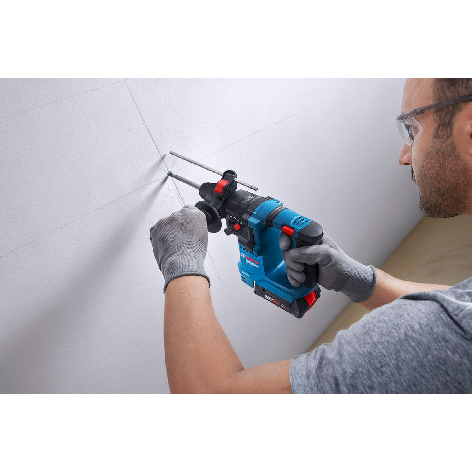Ein Mann bohrt mit einem blau-schwarzen Bohrer in eine weiße Wand. Er trägt eine Schutzbrille und graue Handschuhe.