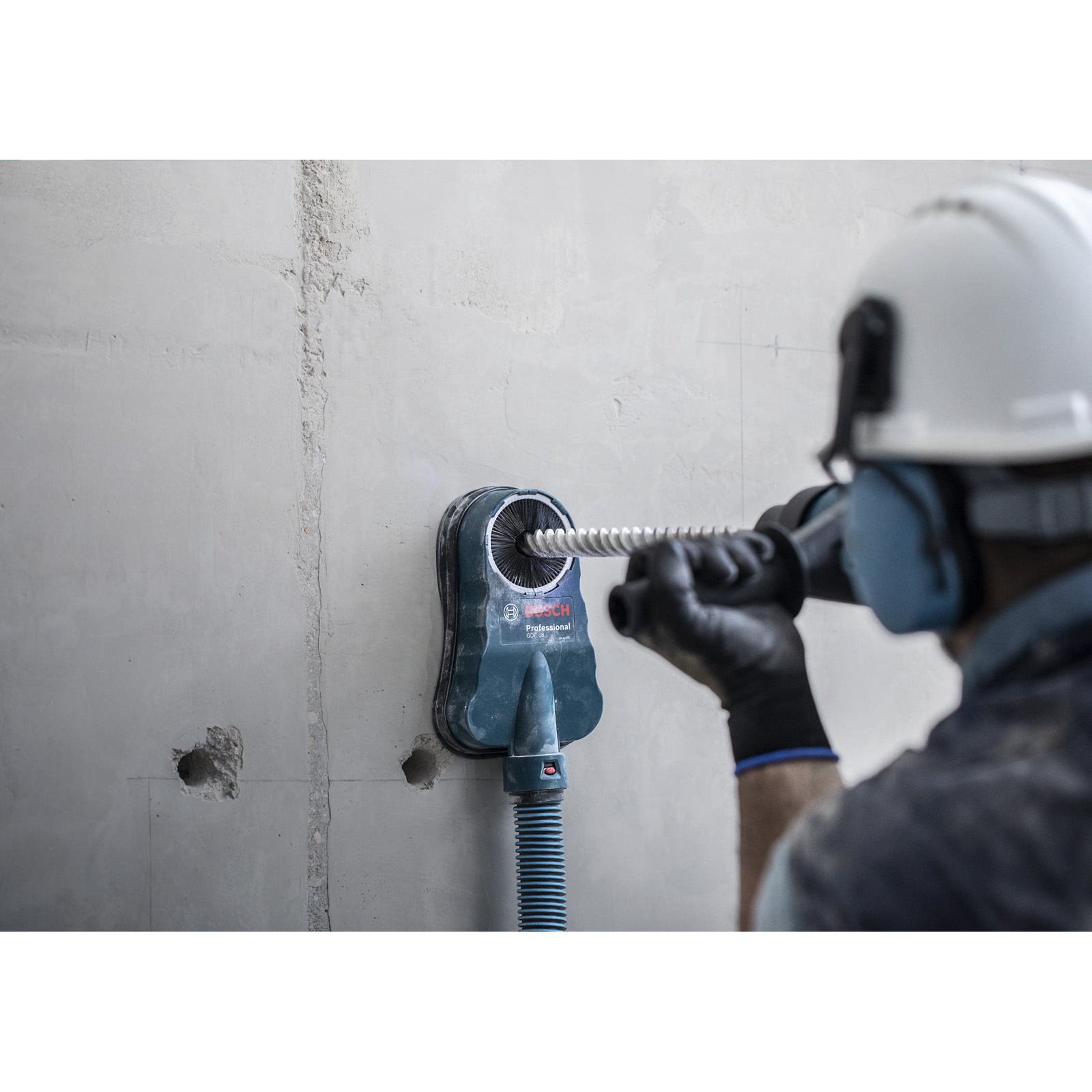 Ein Arbeiter bohrt mit einem Bosch-Werkzeug in die Wand. Er trägt einen weißen Helm und Gehörschutz. Die Wand ist grau mit Löchern.