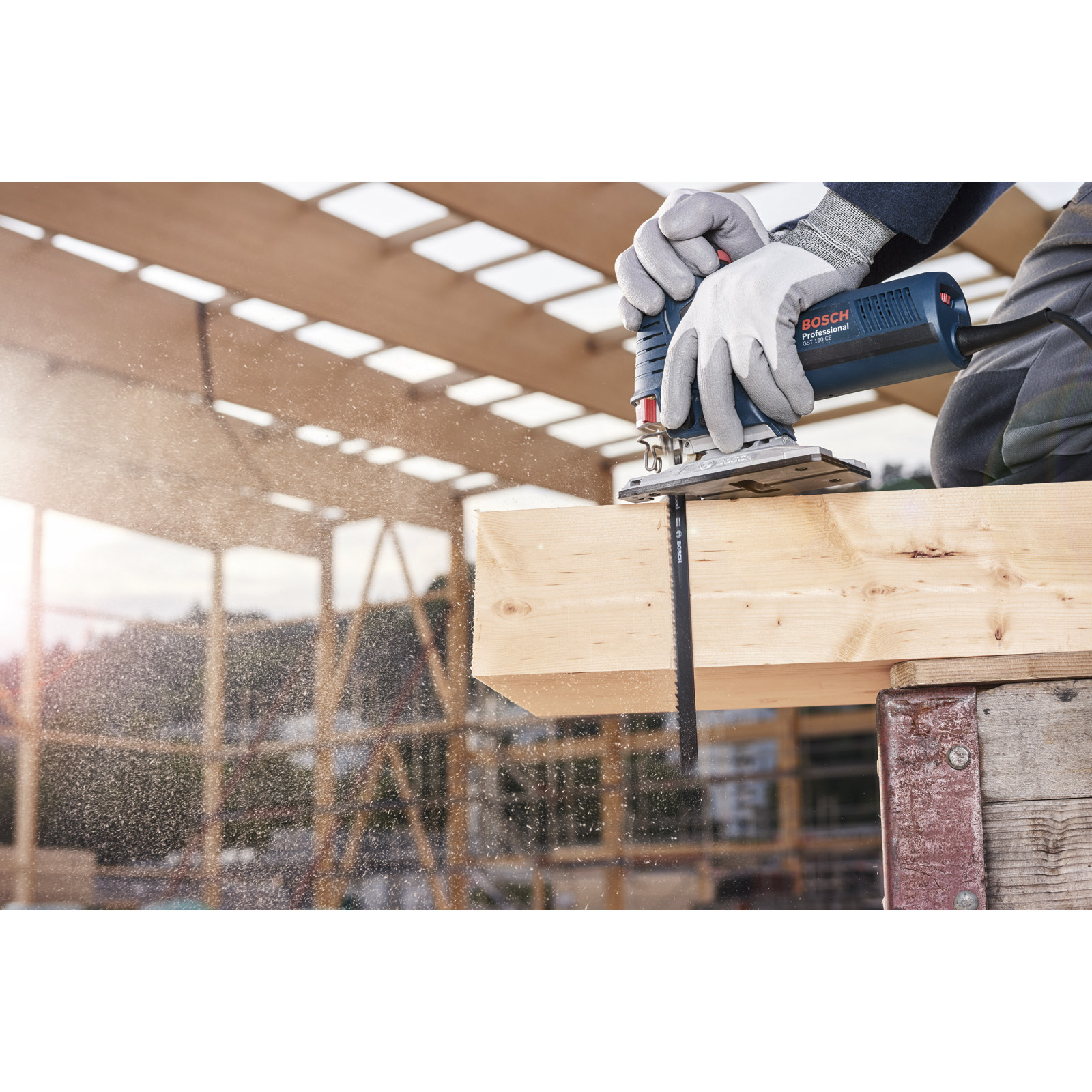 Robotnik używa wyrzynarki Bosch do cięcia drewnianej deski. Wióry lecą. Plac budowy w tle.