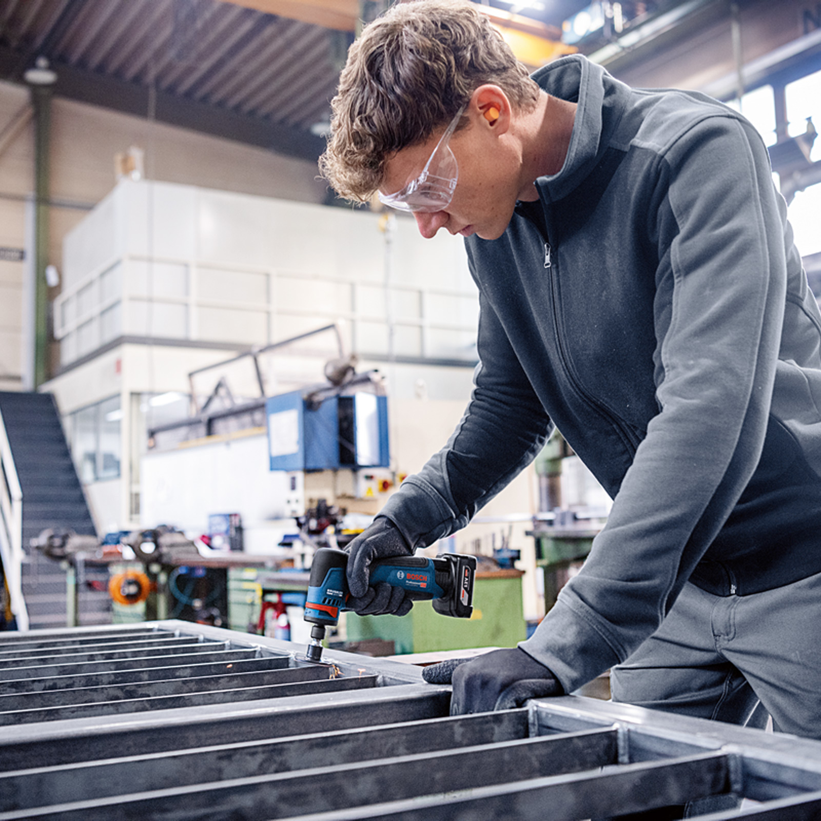 Un homme dans un atelier utilise un outil électrique pour travailler sur un cadre en métal. Il porte des lunettes et des gants.