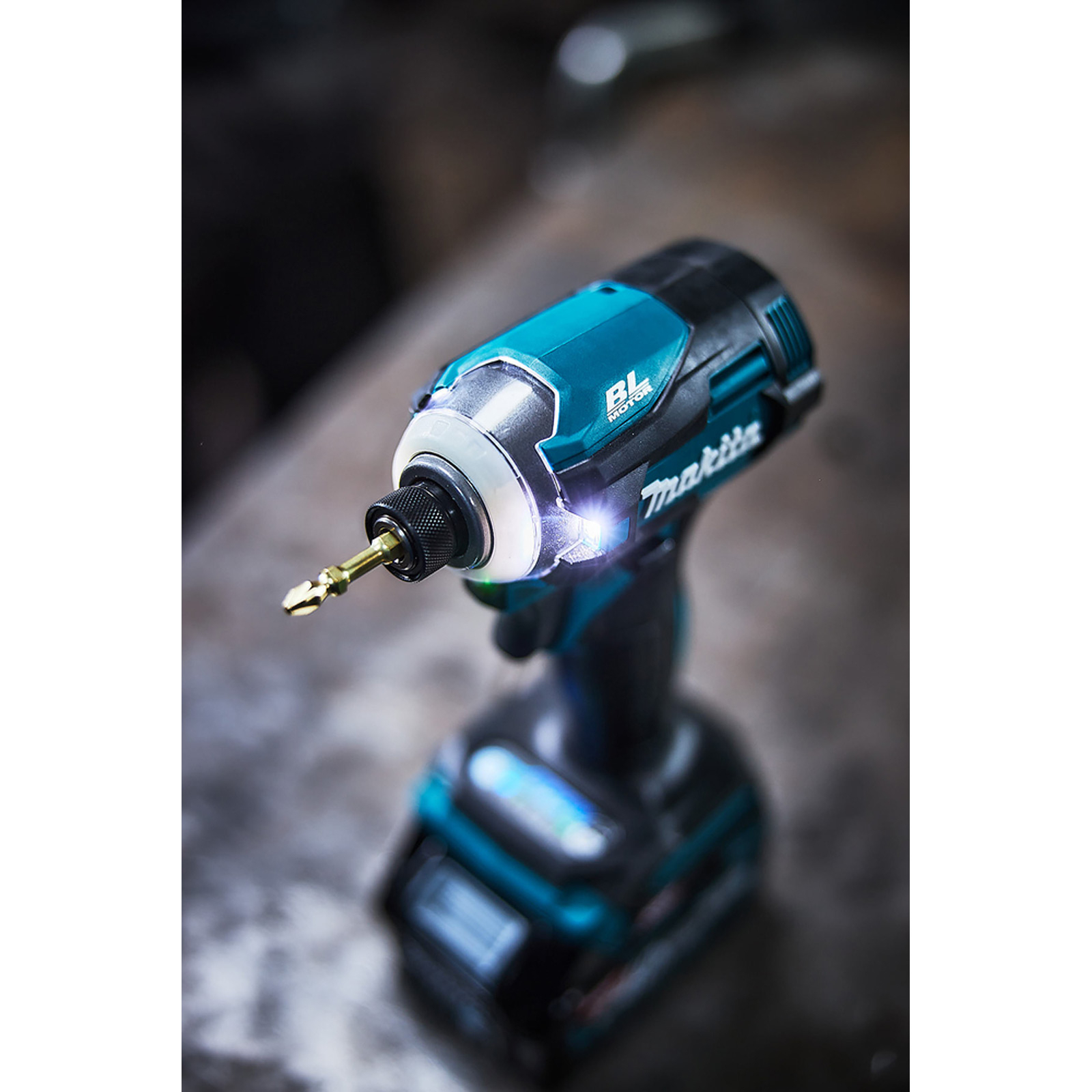 Atornillador de impacto Makita. Combinación de colores turquesa y negro. Una broca dorada adjunta. La herramienta está sobre una superficie gris.