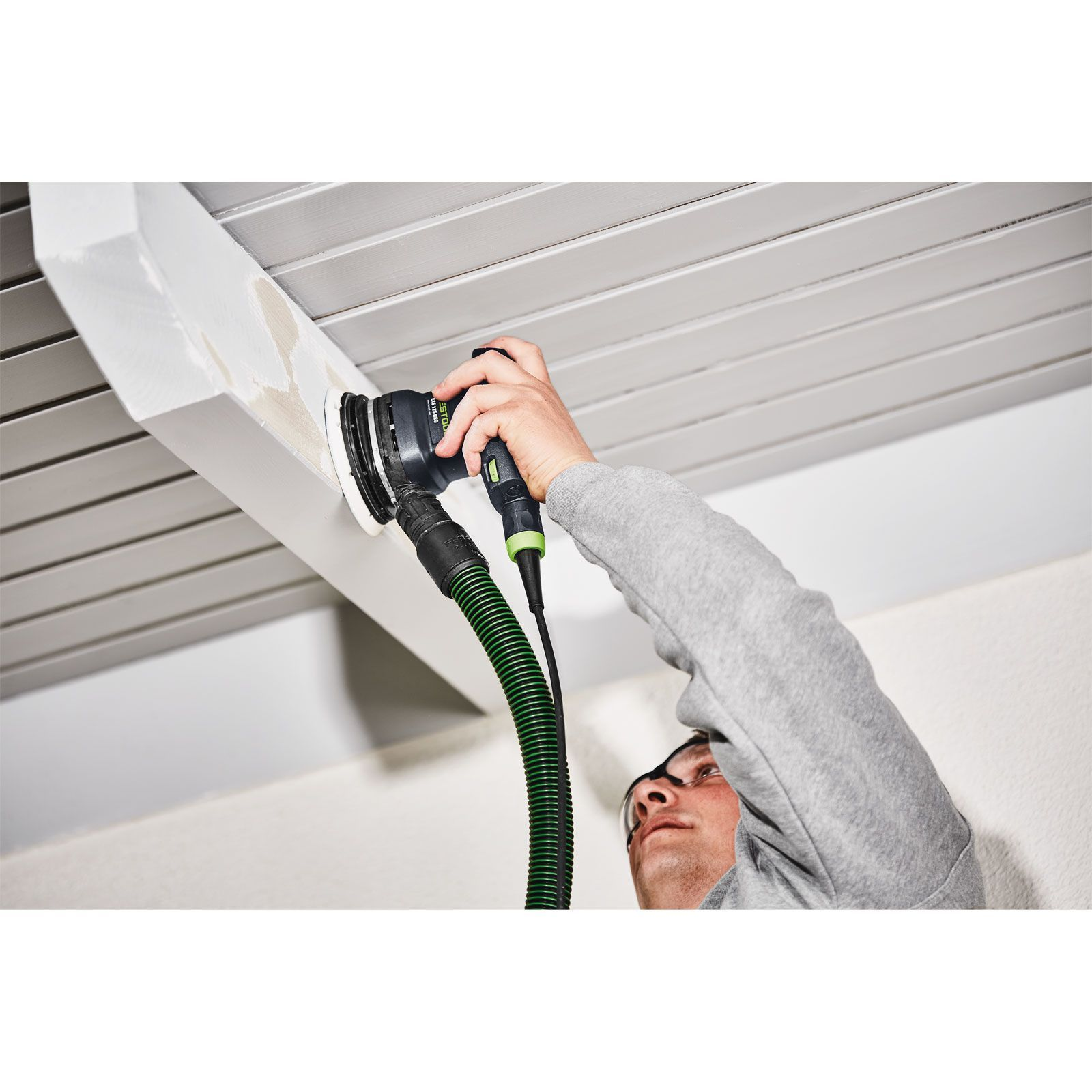 Eine Person schleift einen weißen Holzbalken mit einem Elektroschleifer. Die Person trägt eine Schutzbrille, der Hintergrund ist eine weiße Decke.