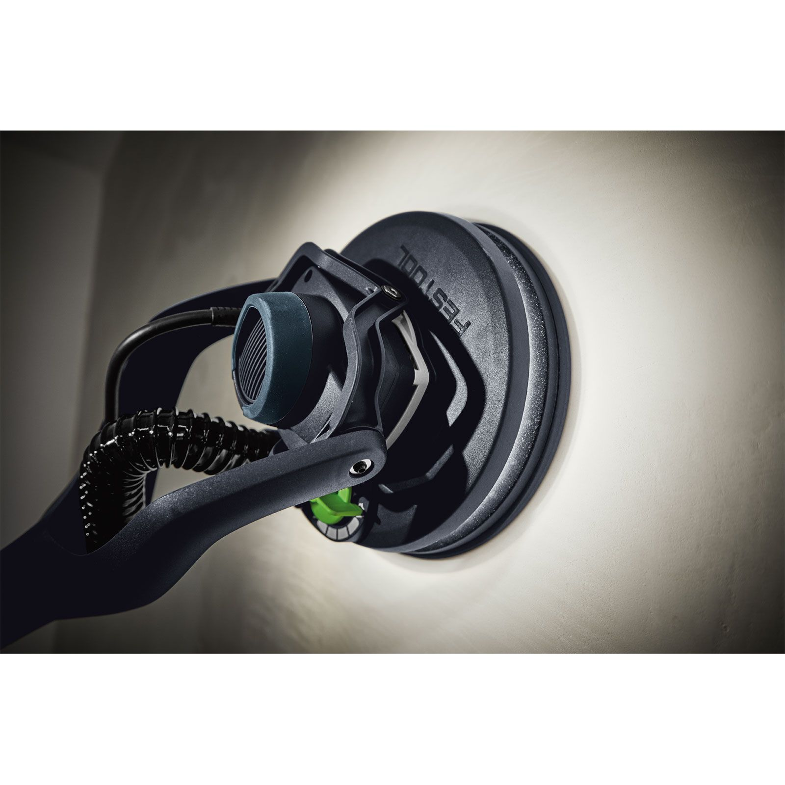 Una lijadora Festool negra está presionada contra una pared de color claro. Tiene una manguera, una palanca verde y una cabeza gris oscura.