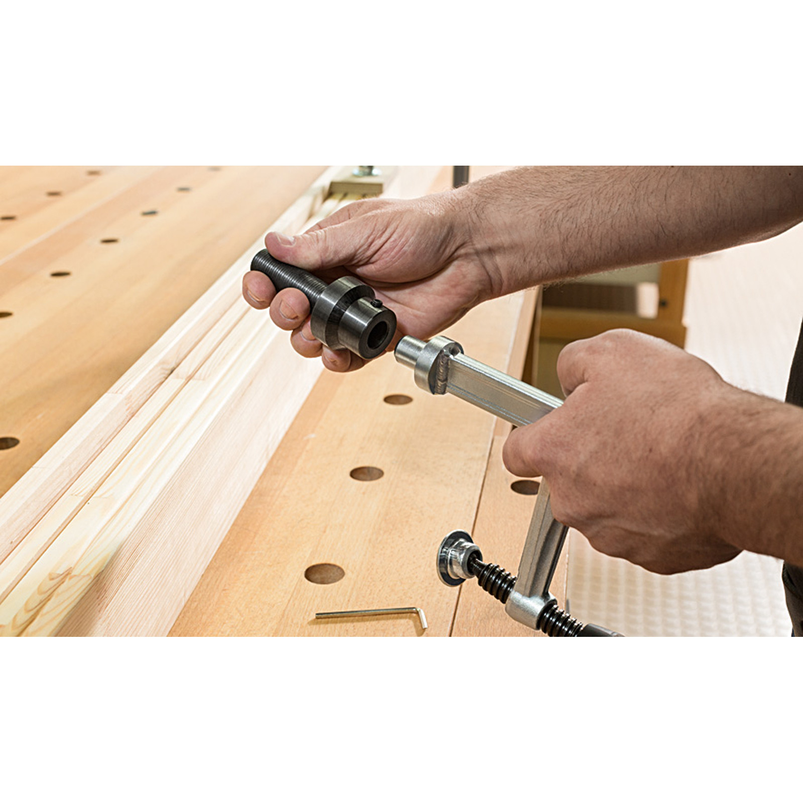 Osoba trzyma zacisk i metalowy cylinder nad drewnianym stołem warsztatowym. Klucz imbusowy leży na stole.