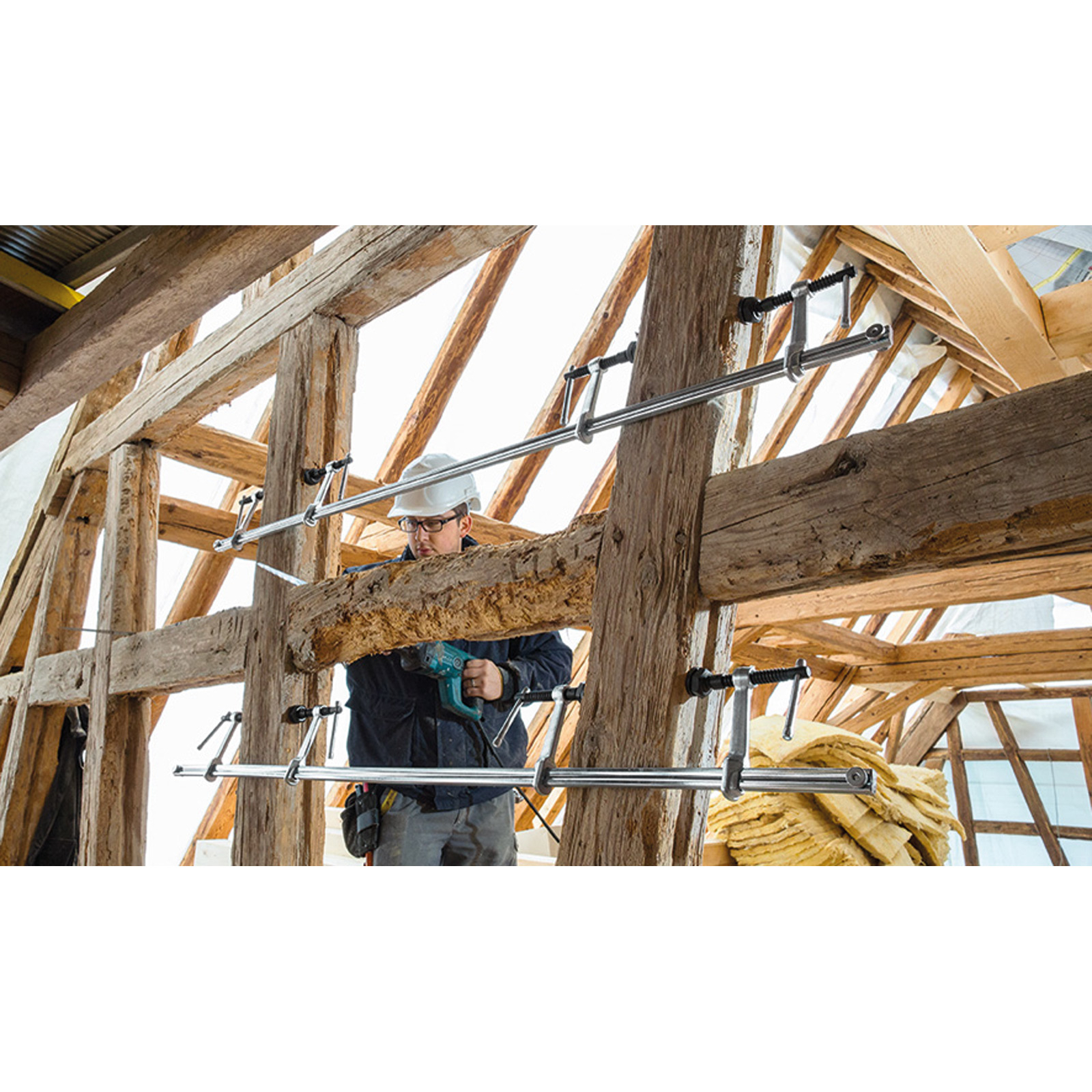 Ein Arbeiter mit Schutzhelm benutzt einen Bohrer auf einem Holzbalken. Klemmen sichern den Balken in einer Holzrahmenkonstruktion.
