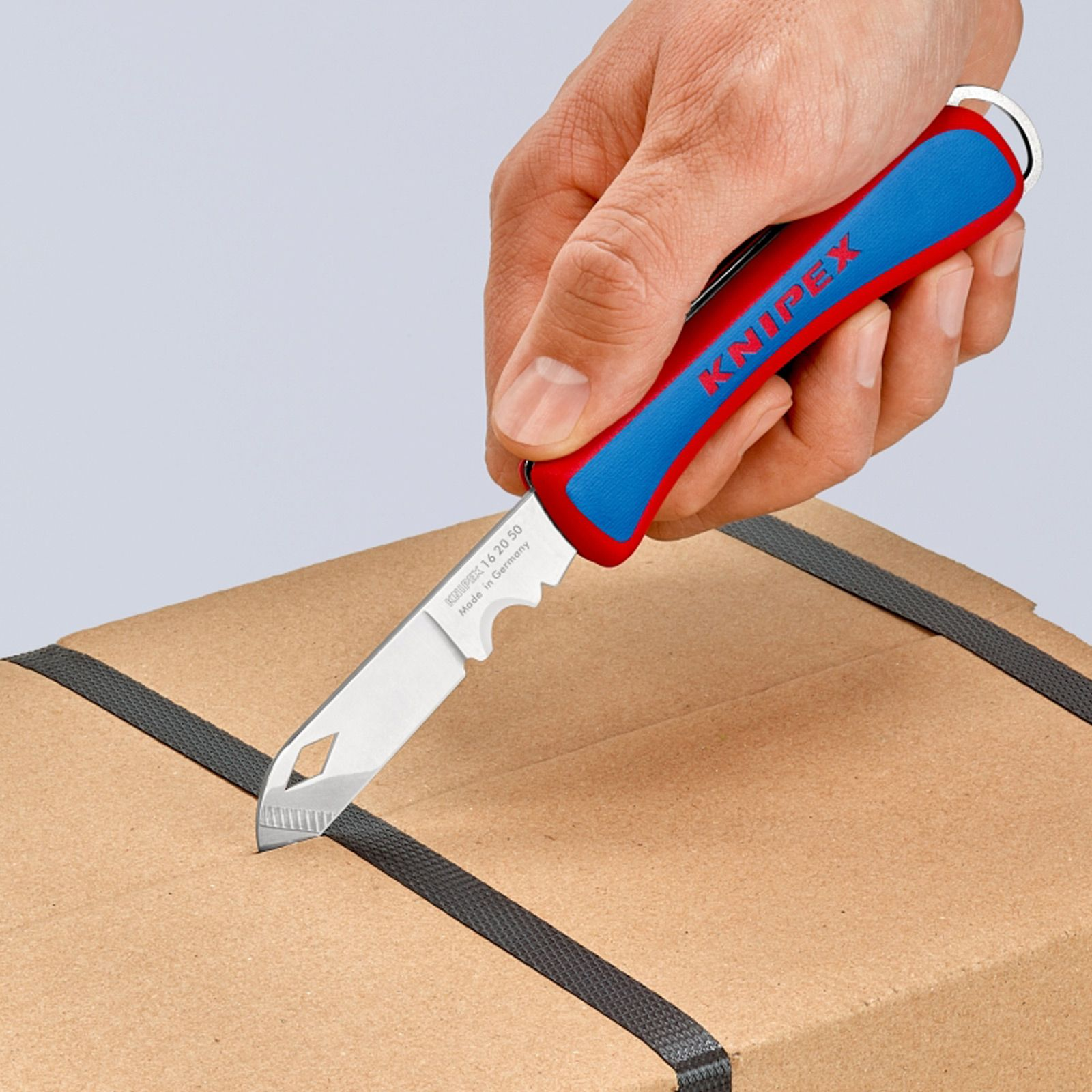 Mano sosteniendo un cuchillo Knipex rojo y azul, cortando una caja de cartón. La caja tiene correas negras.