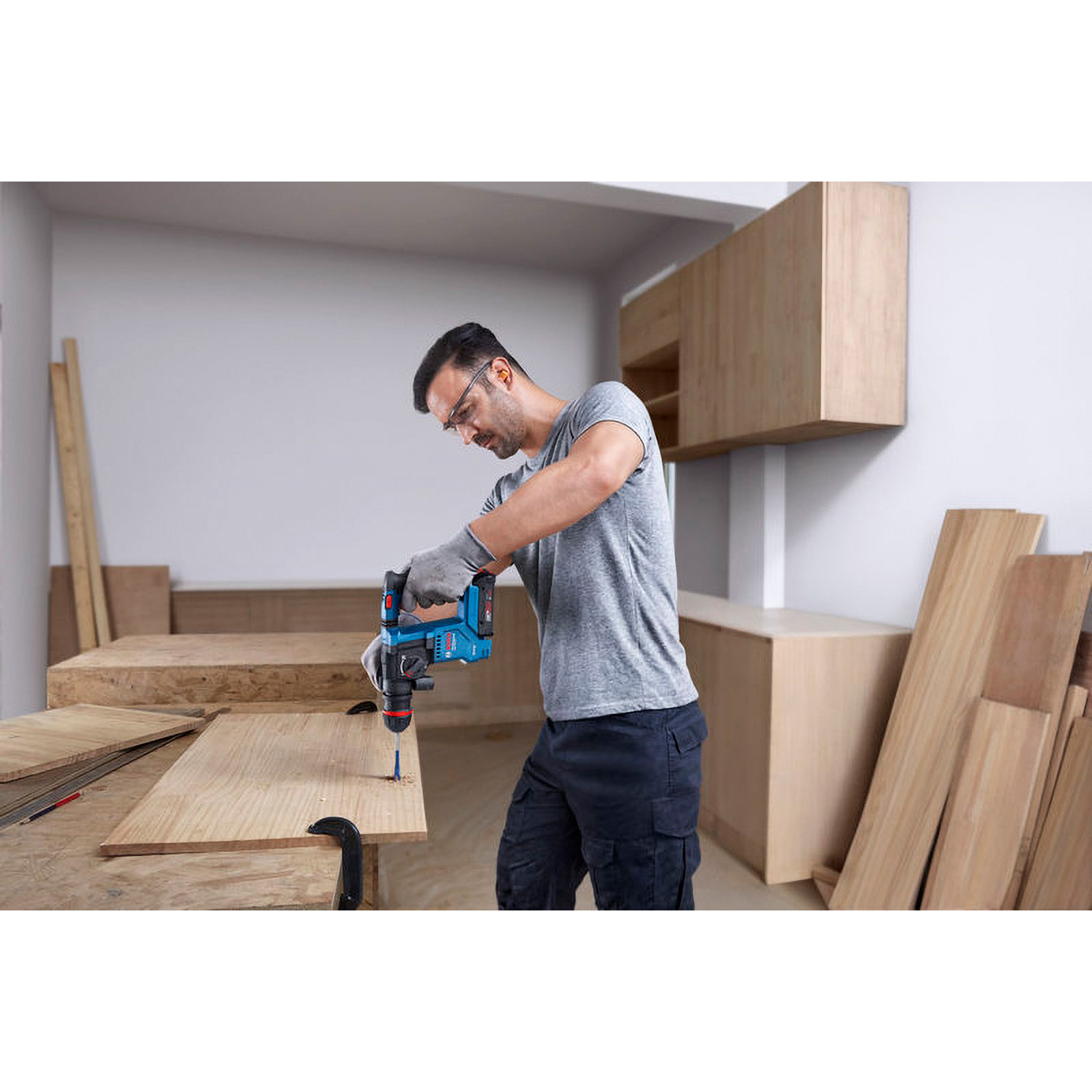 Ein Mann bohrt Holz mit einem blau-schwarzen Bohrer. Er trägt Handschuhe, Brille und ein graues Hemd. Holz und Schränke sind im Hintergrund.