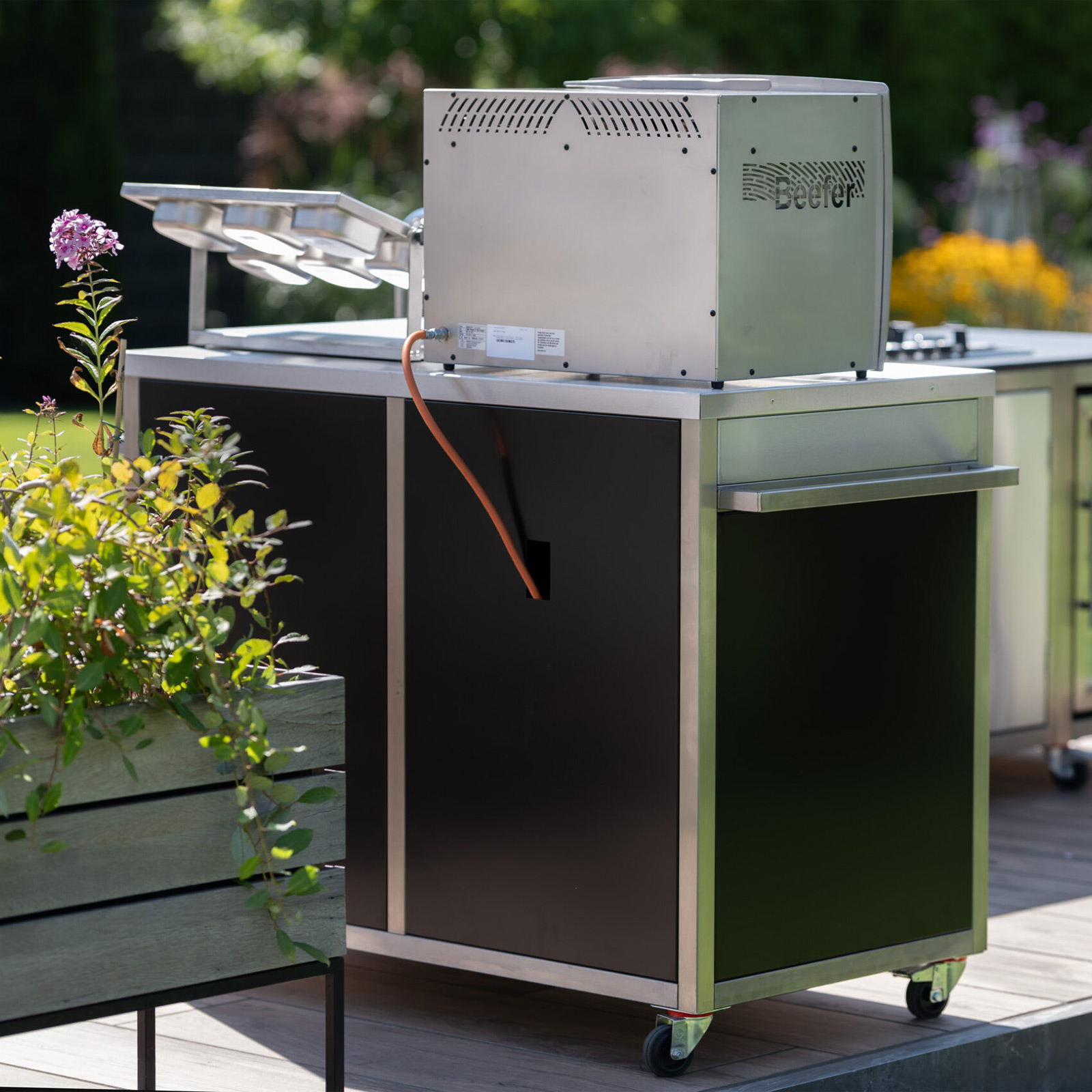 Outdoorküche mit Edelstahlgrill und schwarzem Schrank. Ein Gasschlauch ist angeschlossen. Pflanzen und Blumen sind im Vordergrund.