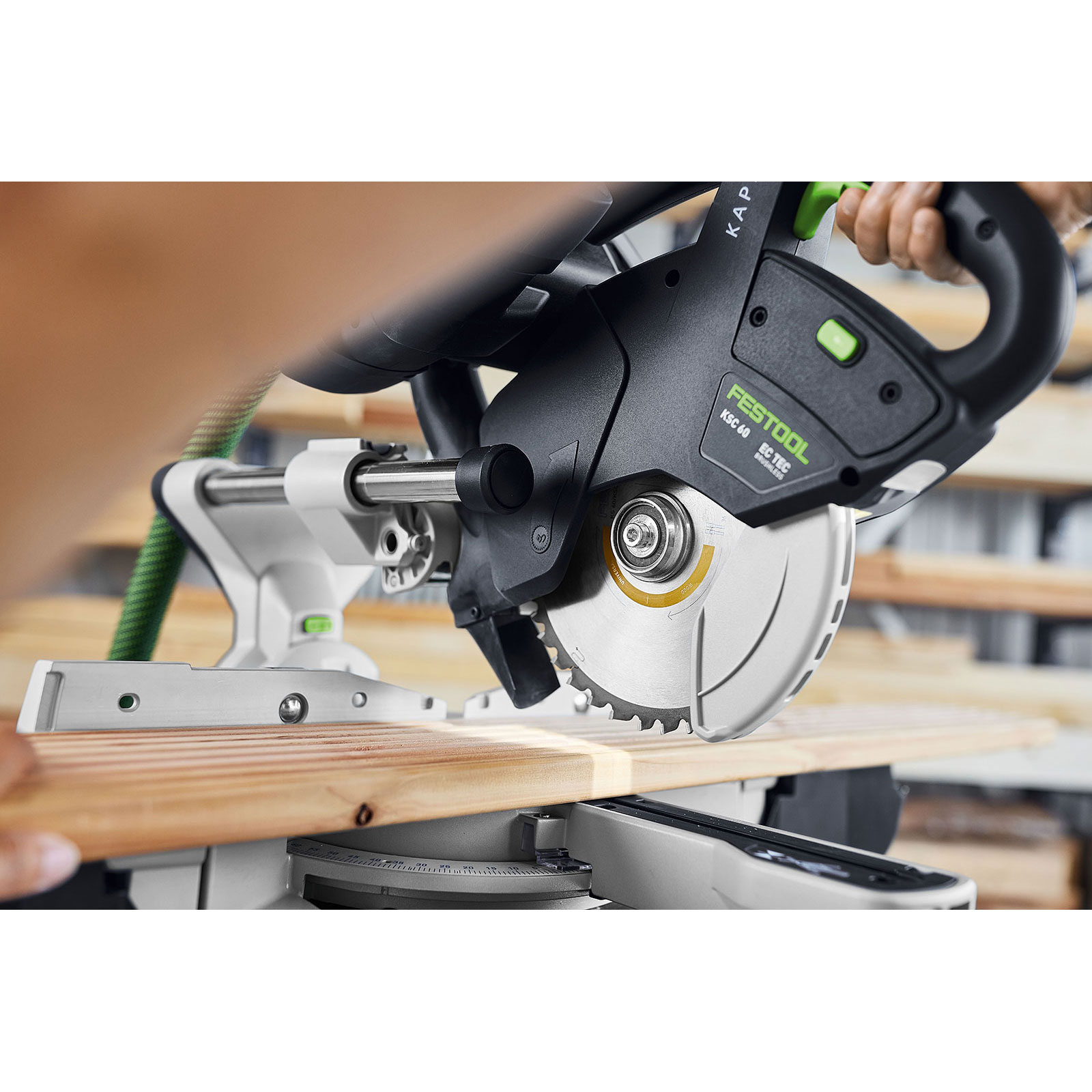 Eine Person benutzt eine Festool KSC 60 Säge, um ein Holzbrett zu schneiden. Die Säge ist schwarz und grün, mit silbernem Blatt.