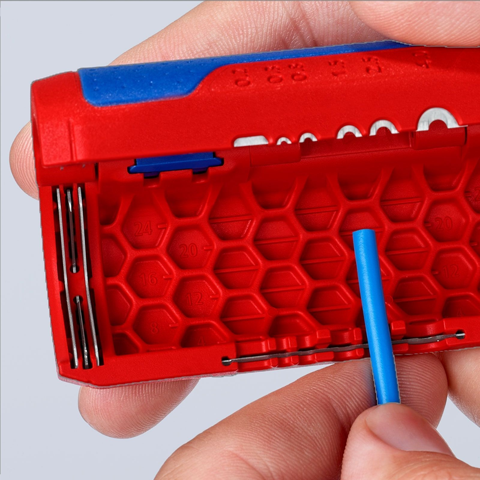 Hand holding a blue wire, inserting it into a red and blue wire stripper. Numbers are visible on the red part.