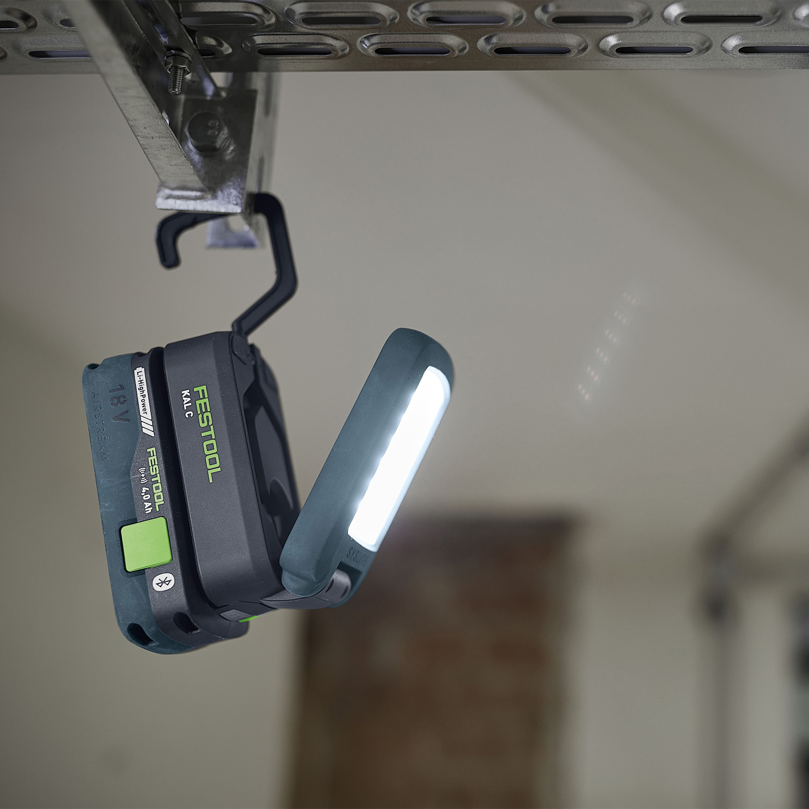 Eine Festool-Arbeitsleuchte ist an einen Metallträger gehängt. Das Licht ist eingeschaltet und nach außen gerichtet. Der Hintergrund ist eine verschwommene Decke.