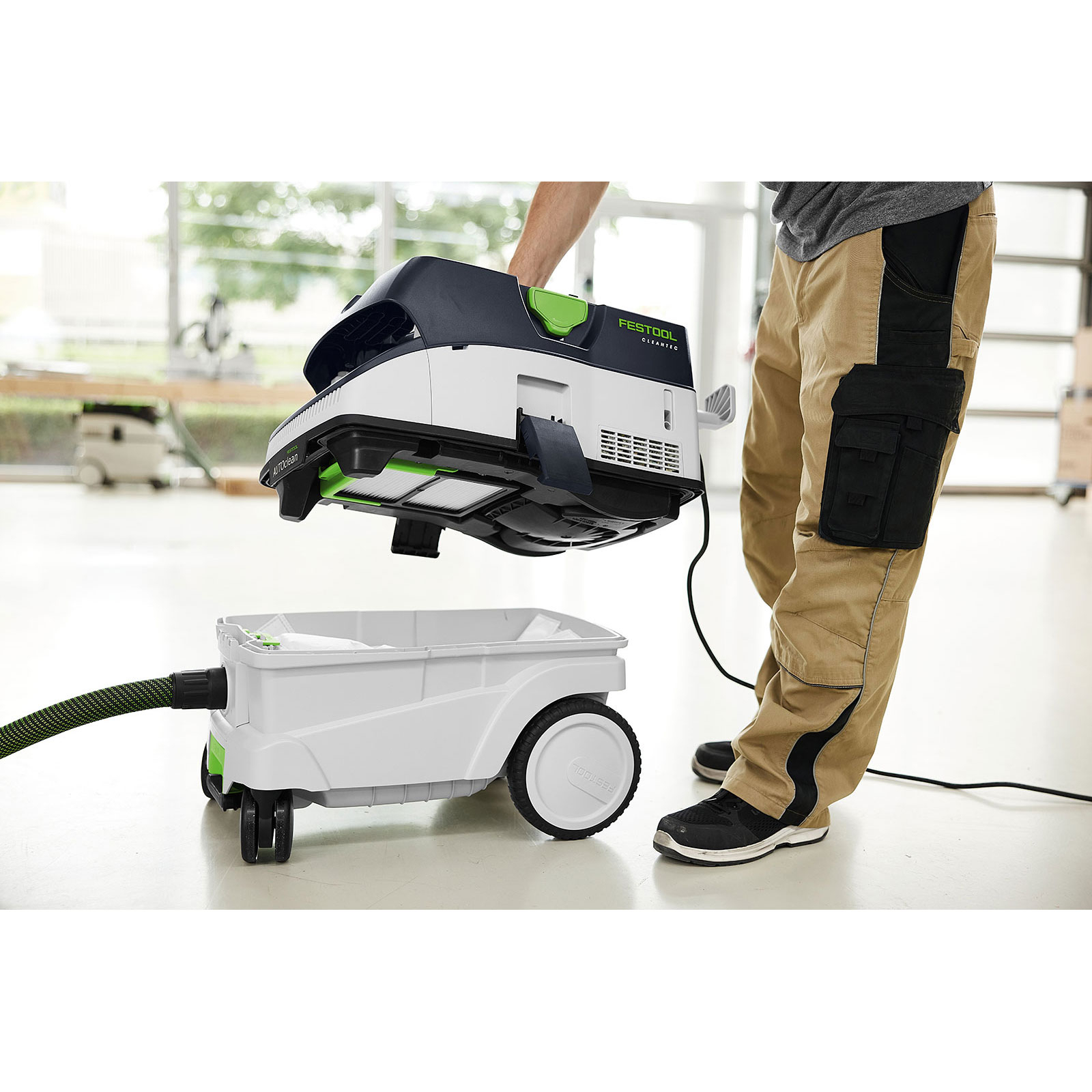 Eine Person hält einen Festool-Staubsauger über seinem Behälter. Der Staubsauger ist schwarz, grün und weiß. Die Person trägt Arbeitshosen.