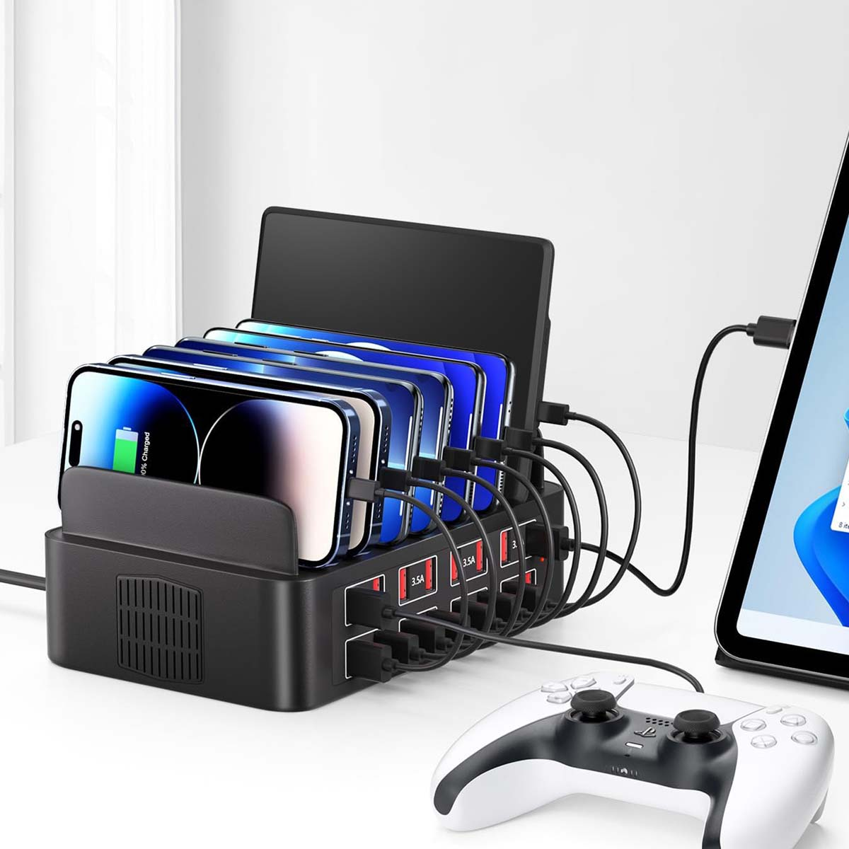 Schwarze Ladestation mit Telefonen, Tablet und Kabeln. Ein weißer Gamecontroller im Vordergrund. Alles auf weißer Oberfläche.