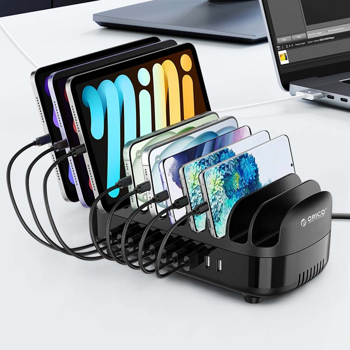 Eine schwarze Ladestation hält mehrere Tablets und Smartphones. Kabel verbinden sie mit der Station. Ein Laptop befindet sich im Hintergrund.