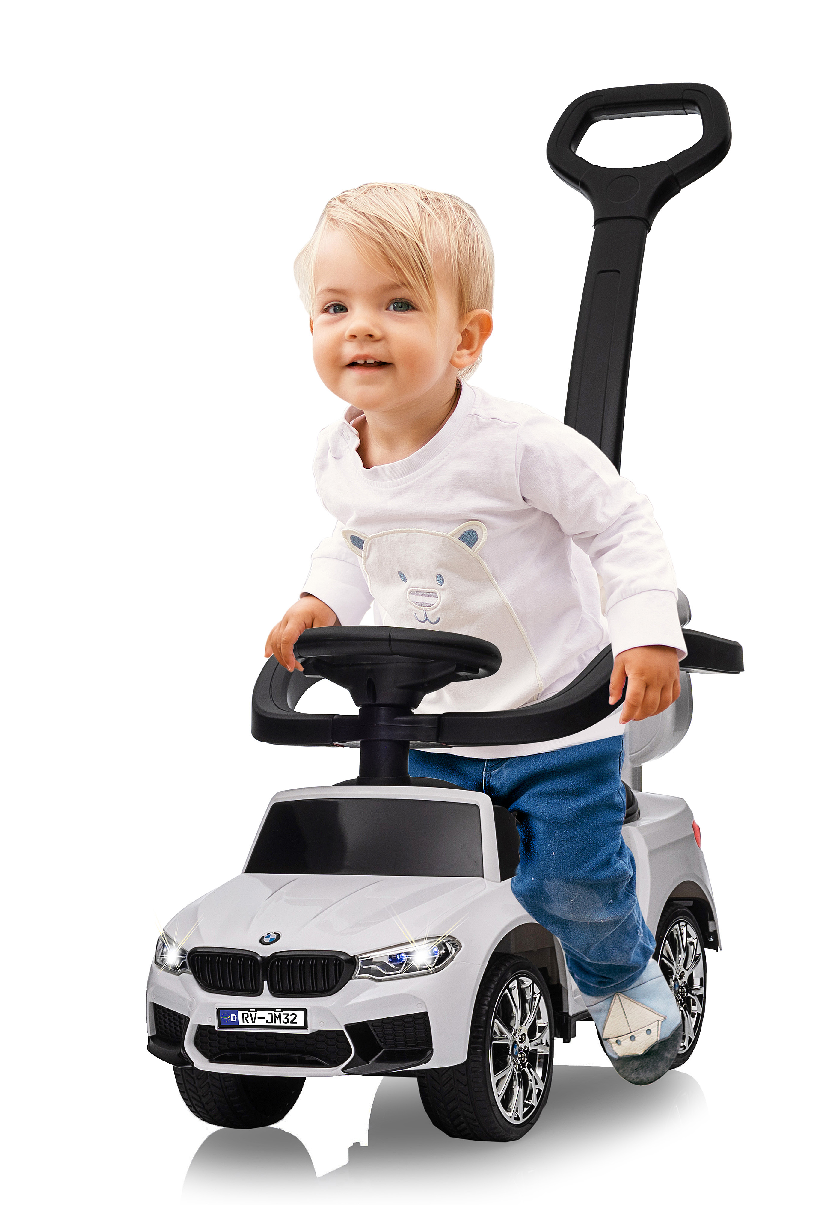 Un bambino piccolo è seduto in un'auto giocattolo bianca, sorridendo. L'auto ha un manico. Il bambino indossa jeans blu e una camicia bianca.