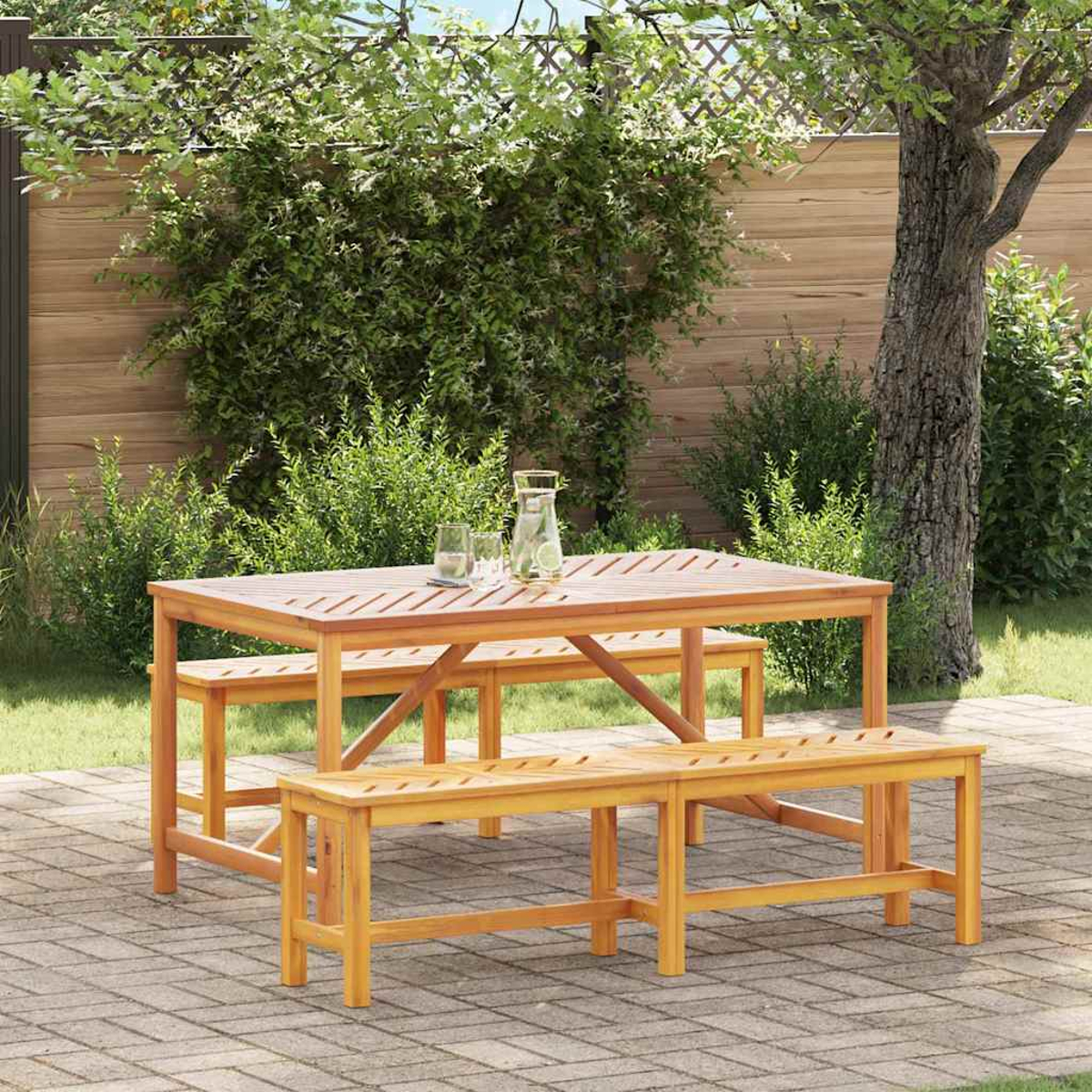 Juego de comedor de madera al aire libre en un patio de ladrillos. Incluye mesa, bancos, jarra y vasos, con follaje verde al fondo.